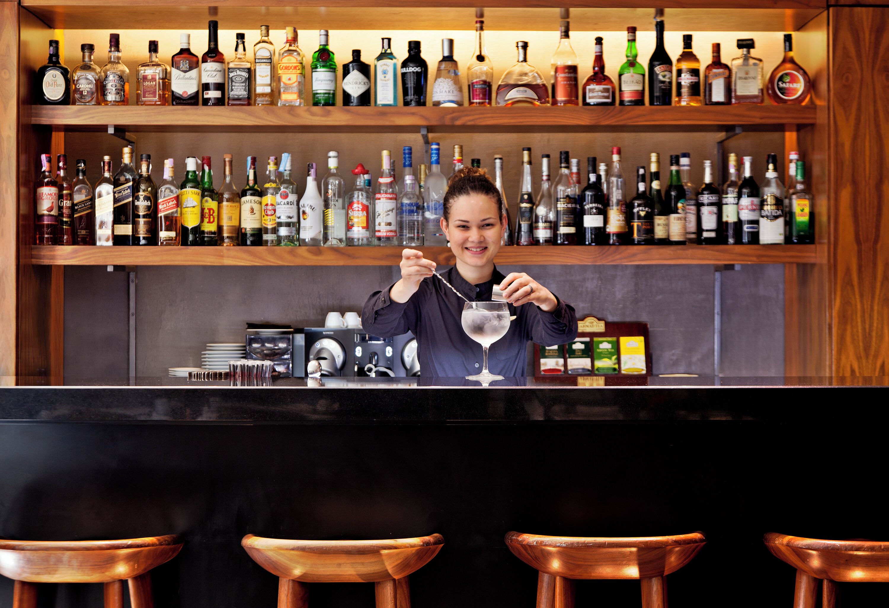 Una zona de bar con un bartender, estantes llenos de botellas de licor y taburetes de madera.