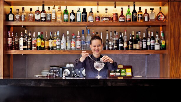A bar area with a bartender, shelves of liquor bottles, and wooden stools.
