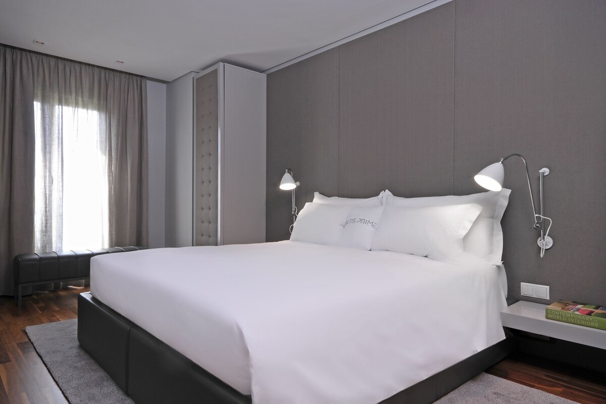 Un dormitorio moderno con una cama grande, dos lámparas de noche, un banco y un libro en la mesita de noche.