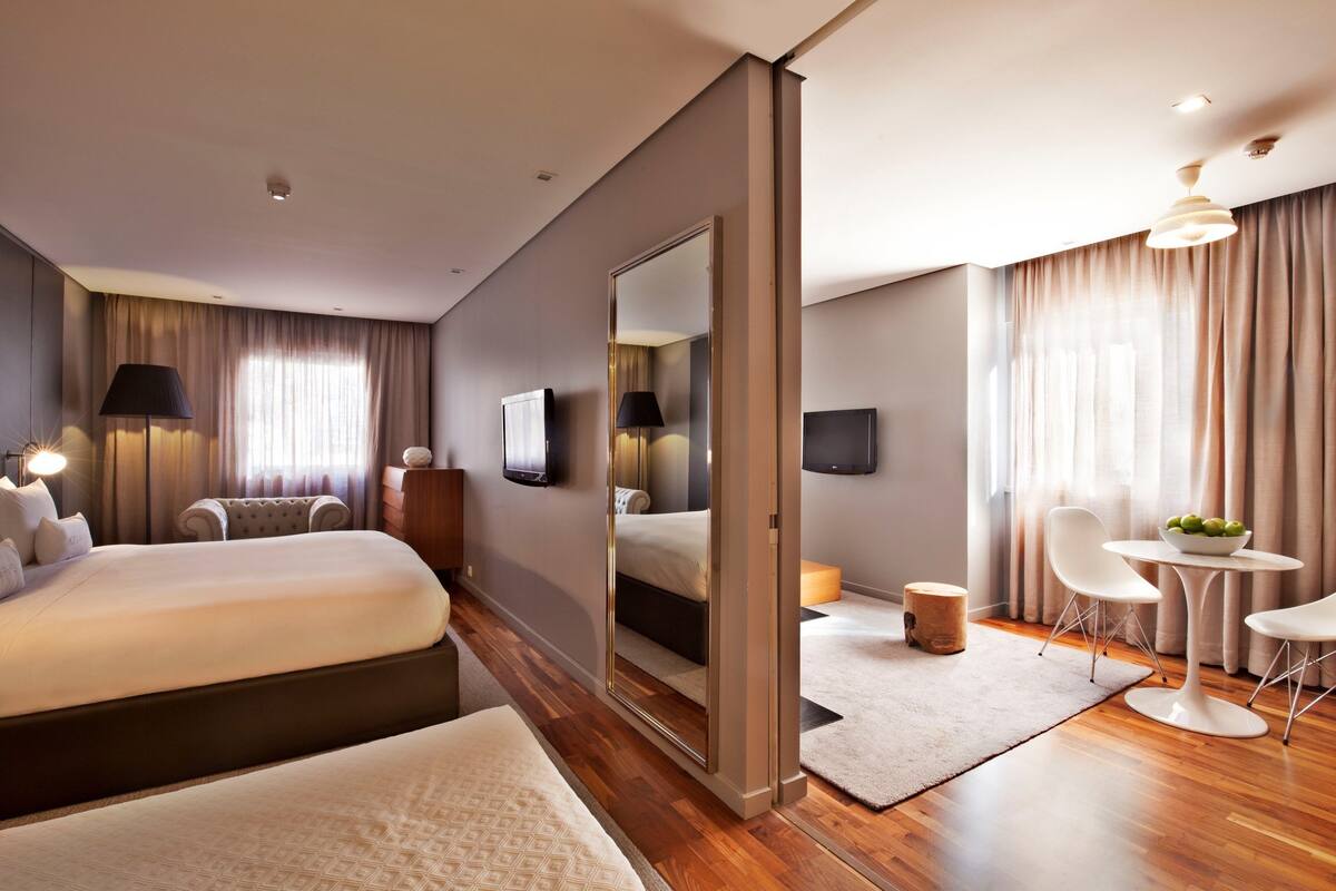 Ein modernes Hotelzimmer mit zwei Betten, einer Sitzecke inklusive Tisch und Stühlen, einem großen Spiegel und einem Flachbildfernseher.