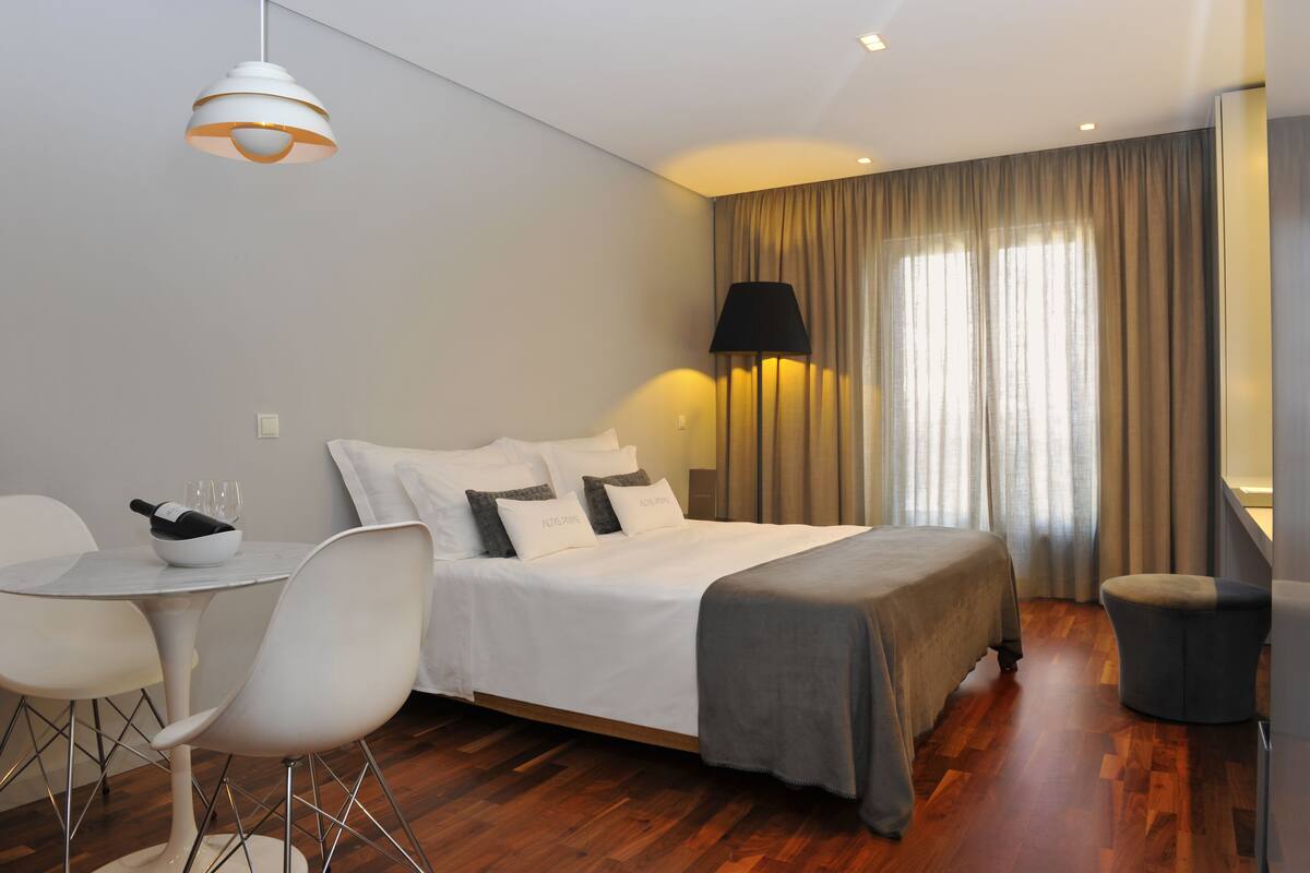 Ein modernes Hotelzimmer mit einem ordentlich bezogenen Bett, einem Esstisch mit Stühlen, einem kleinen runden Tisch mit Schüssel und Gläsern sowie einem großen Fenster mit Vorhängen.