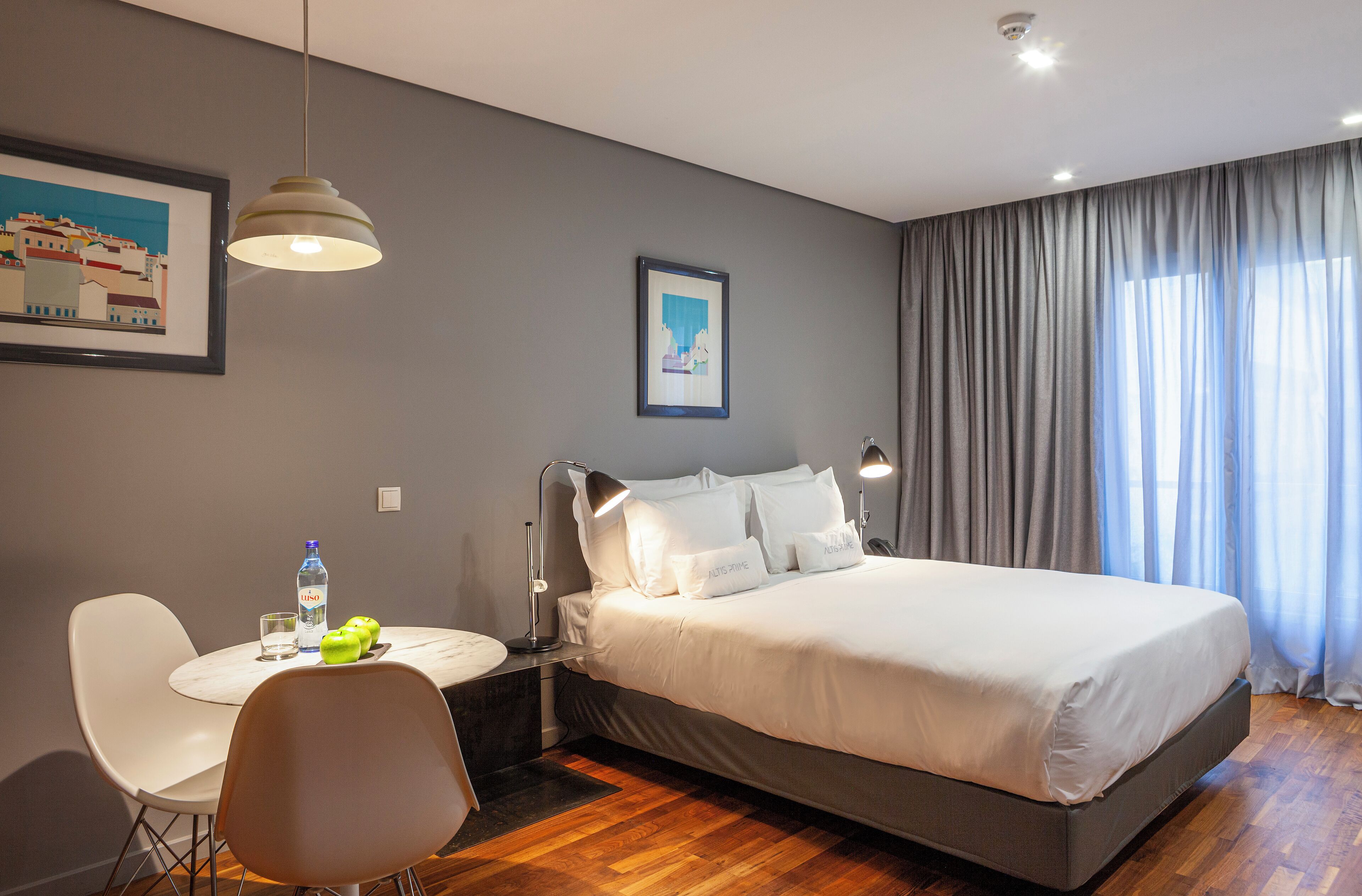 A modern hotel room with a bed, bedside lamps, a desk with a chair, a framed picture on the wall, and a window with curtains.