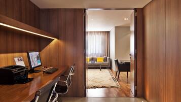 Een moderne hotelkamer met een houten bureau, computer en printer, een zithoek met een bank en salontafel, en uitzicht op de woonkamer.