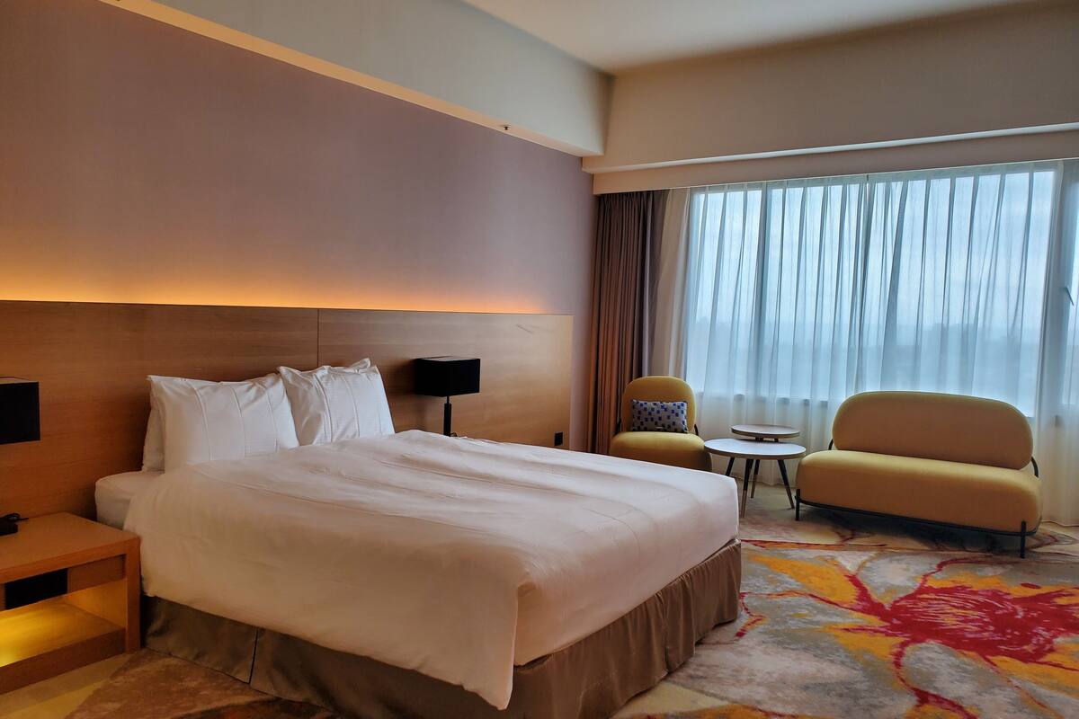 Een hotelkamer met een groot bed, een nachtkastje, een lamp, een klein rond tafeltje, een gele fauteuil en een raam met dunne gordijnen.