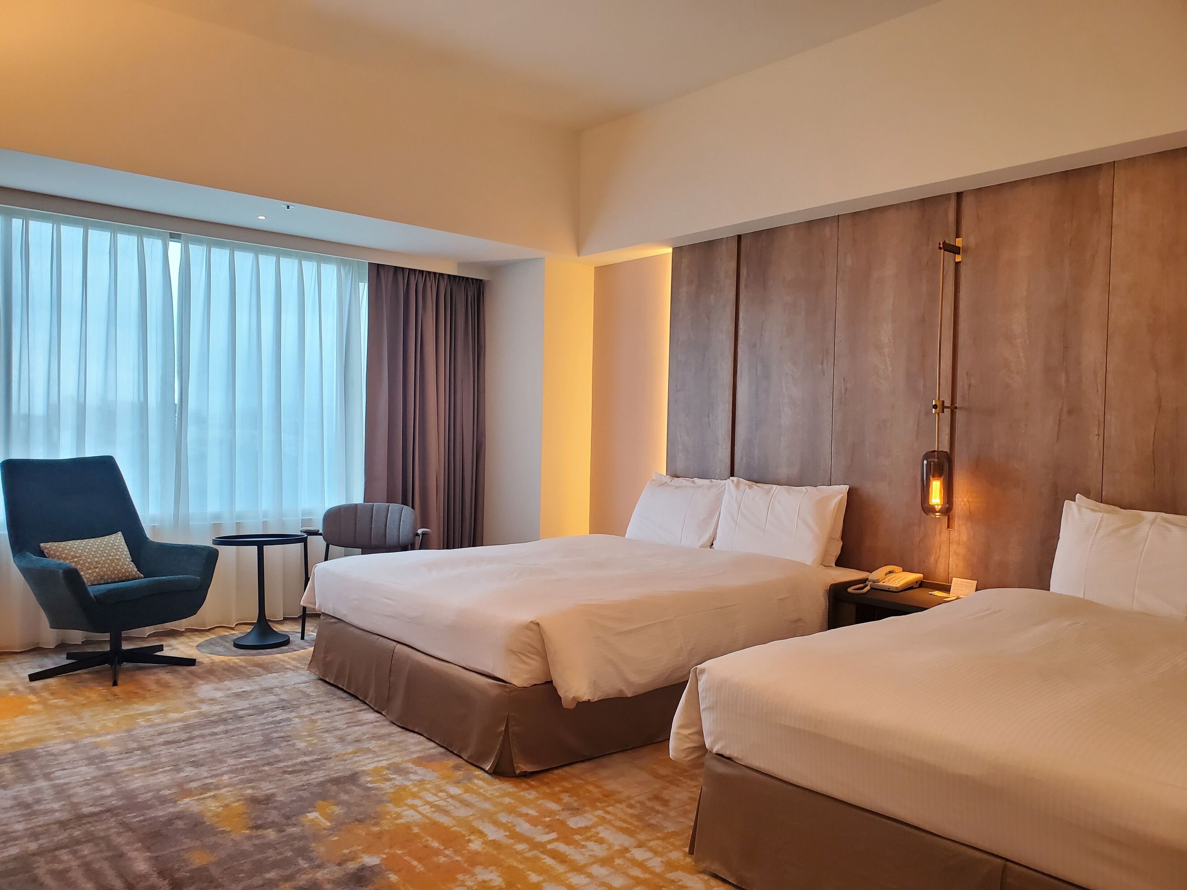 A hotel room with two beds, a chair, a small table, and a large window with curtains.