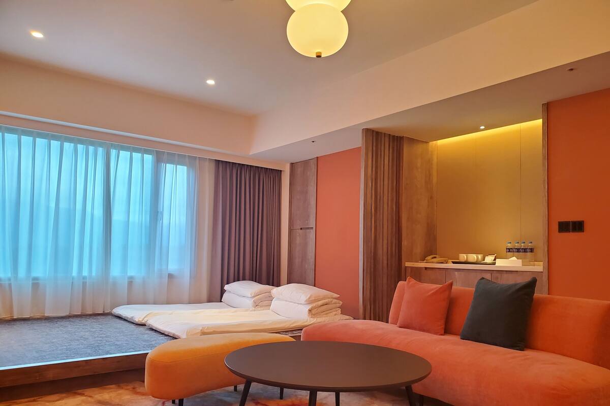Een moderne hotelkamer met een bed, een bank, een klein tafeltje en een groot raam met gordijnen.