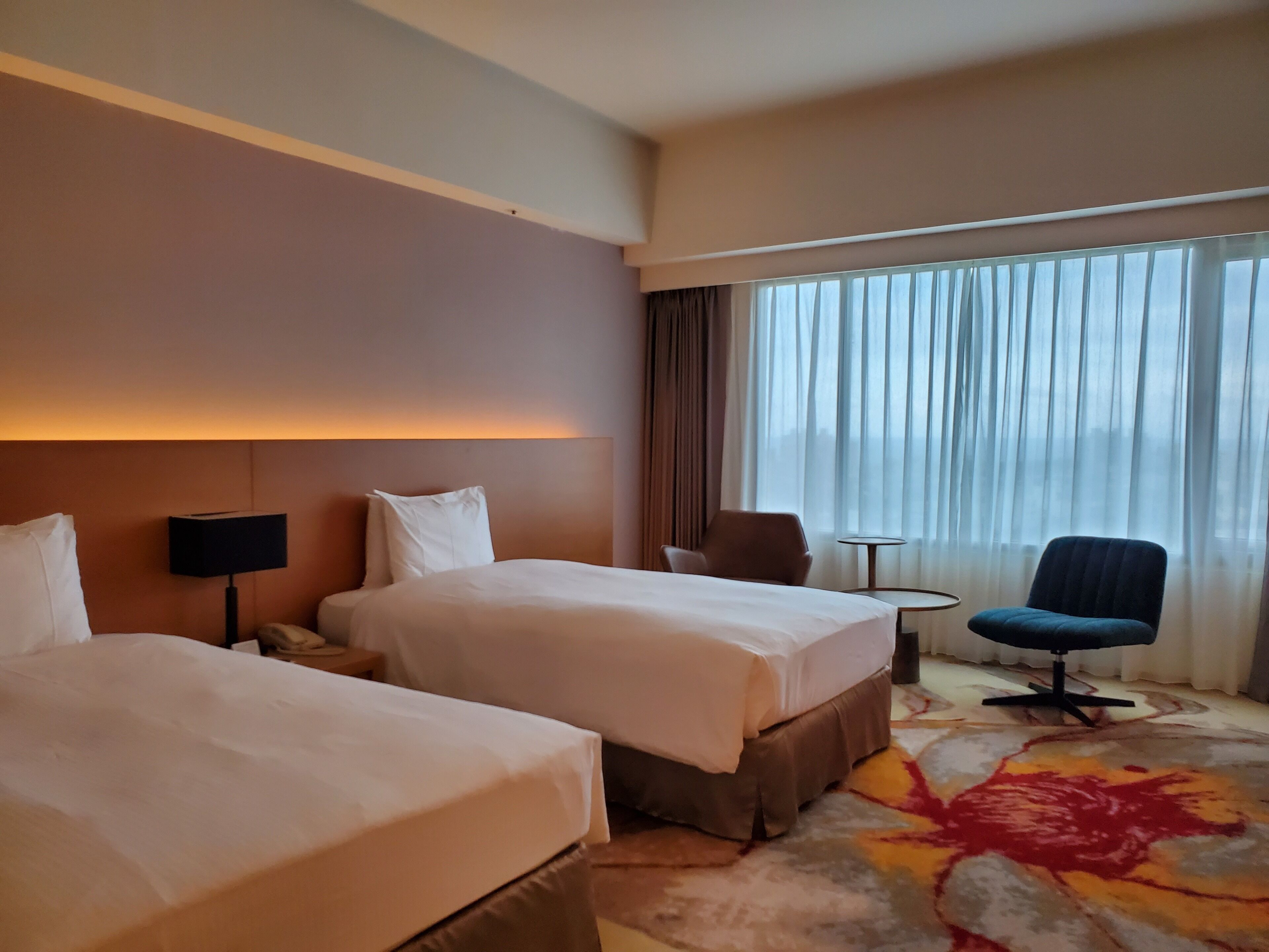 A hotel room with two beds, a desk, a chair, and a large window with curtains.