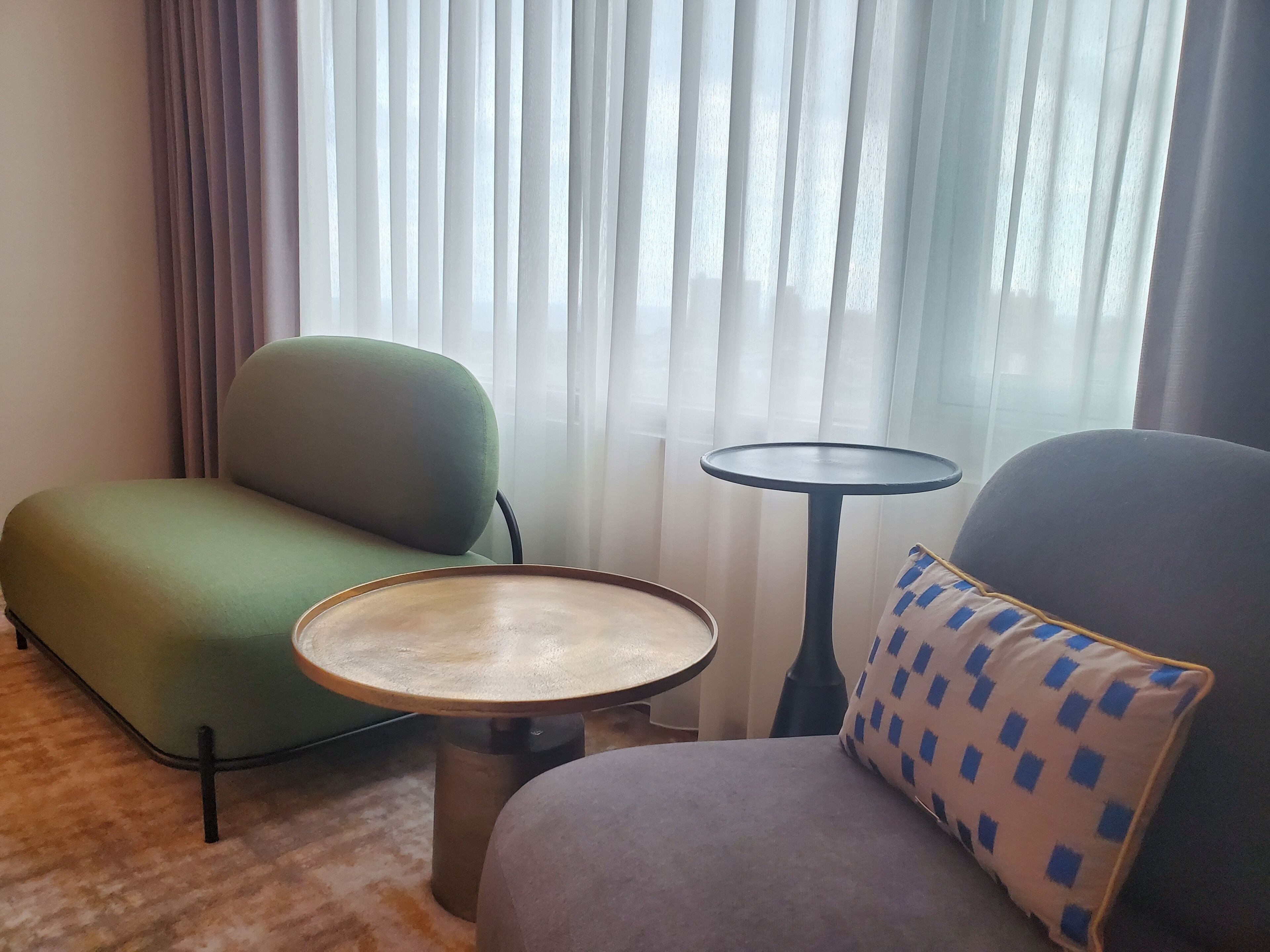 A room with a green armchair, a grey armchair, a small round table, and a window with sheer curtains.