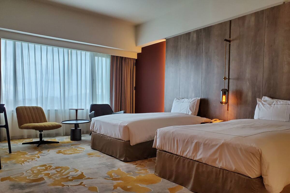 Een hotelkamer met twee bedden, een bank, een stoel, een klein tafeltje en een groot raam met gordijnen.