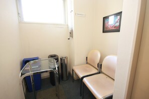 Una habitación pequeña y limpia con una única silla color beige, una mesa de vidrio, un cubo de basura azul y un cubo de basura plateado.
