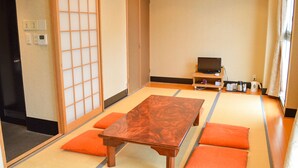 Une pièce de style japonais traditionnel, avec un sol recouvert de tatamis, une basse table en bois, des coussins orange et un téléviseur posé sur un meuble.