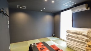Une pièce avec un sol recouvert de tatamis, un lit avec une literie blanche et orange, une pile d’oreillers et un appareil de climatisation.