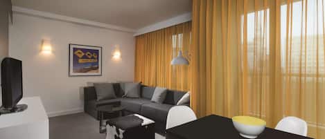 A modern living room with a sectional sofa, a dining table with chairs, a flat-screen TV on a white stand, and a framed picture on the wall.