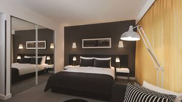 Una habitación de hotel moderna con una cama grande, mesitas de noche con lámparas, un espejo y un amplio ventanal con cortinas.