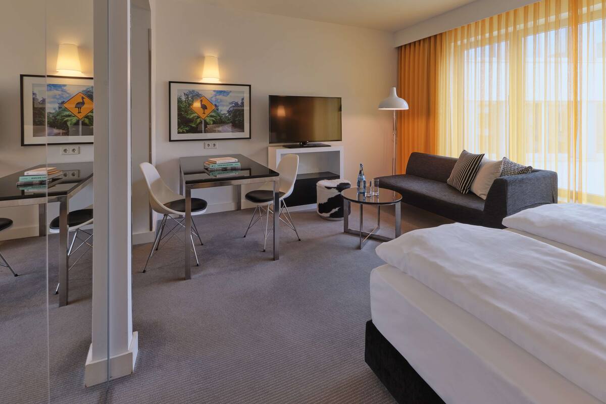 Habitació d'hotel amb un llit, un sofà, una taula de treball amb cadires, un televisor i obres d'art a les parets.