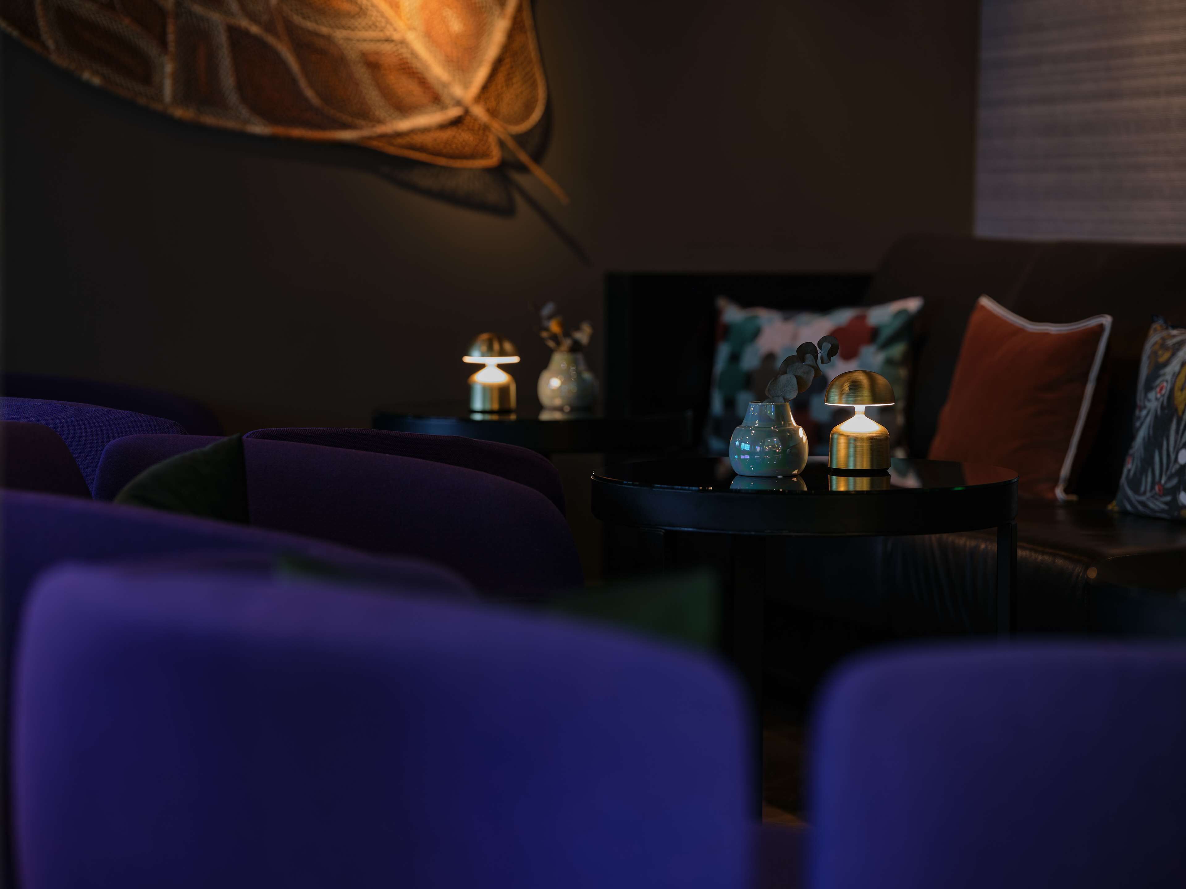 Ein schwach beleuchteter Raum mit violetten Sitzgelegenheiten, einem kleinen Tisch mit zwei Lampen und einer dekorativen Vase.
