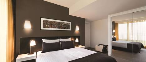 Una habitación de hotel moderna con una cama grande, dos mesitas de noche con lámparas, un espejo amplio y una obra de arte enmarcada en la pared.