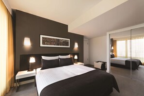 Une chambre d’hôtel moderne, dotée d’un grand lit, de deux tables de chevet avec des lampes, d’un grand miroir et d’une œuvre d’art encadrée au mur.