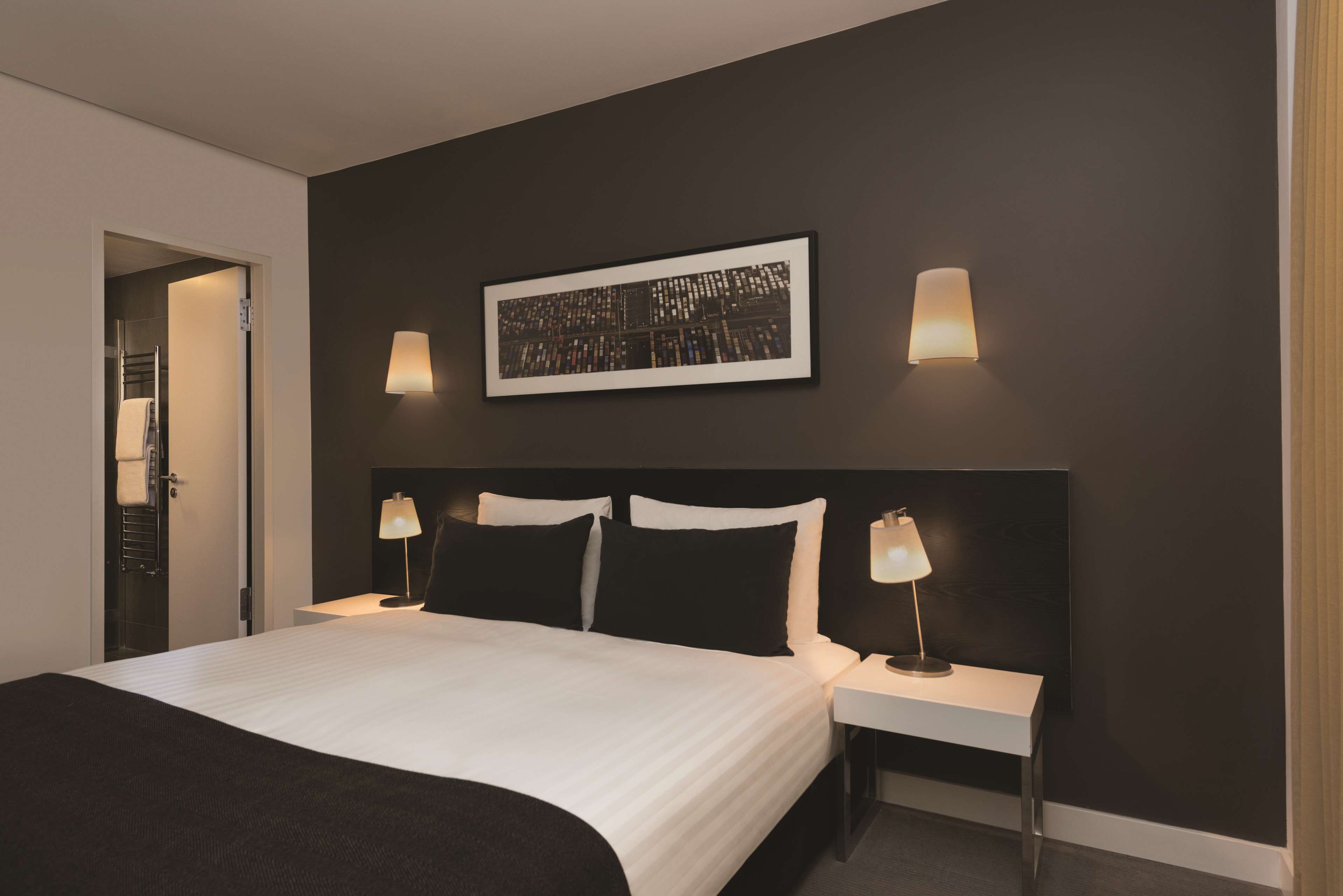 Quarto de hotel moderno com uma cama grande, duas mesas de cabeceira com luminárias, uma obra de arte emoldurada na parede e uma parede de cor escura.
