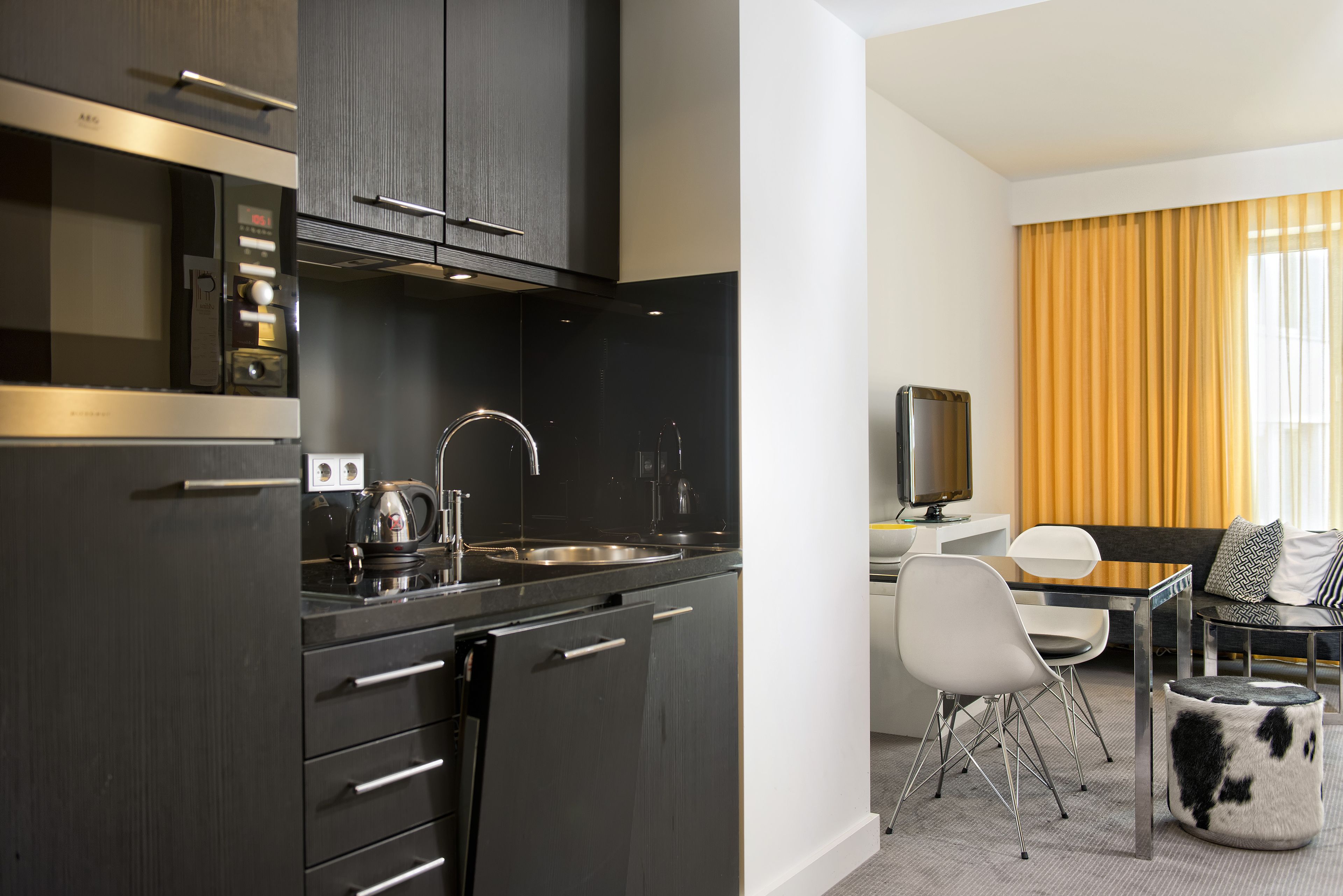 Une cuisine moderne avec des meubles de couleur sombre, un évier en acier inoxydable et une machine à café. On y trouve également un coin repas avec une table blanche, des chaises et un canapé agrémenté de coussins à motifs.