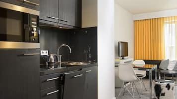 A modern kitchen with dark cabinetry, a stainless steel sink, and a coffee maker. There is a dining area with a white table, chairs, and a sofa with patterned cushions.