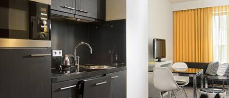 Une cuisine moderne avec des meubles de couleur sombre, un évier en acier inoxydable et une machine à café. On y trouve également un coin repas avec une table blanche, des chaises et un canapé agrémenté de coussins à motifs.