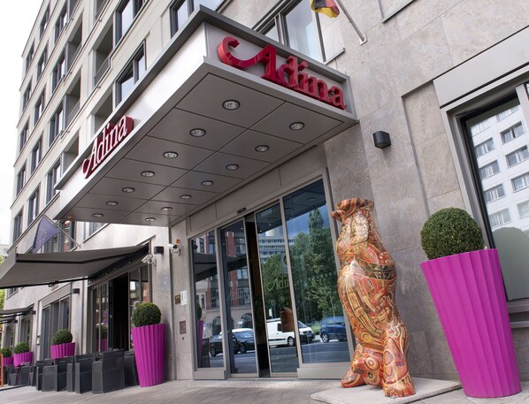L’entrée d’un hôtel, avec une façade moderne, de grandes portes vitrées et une enseigne « Adina » bien visible.