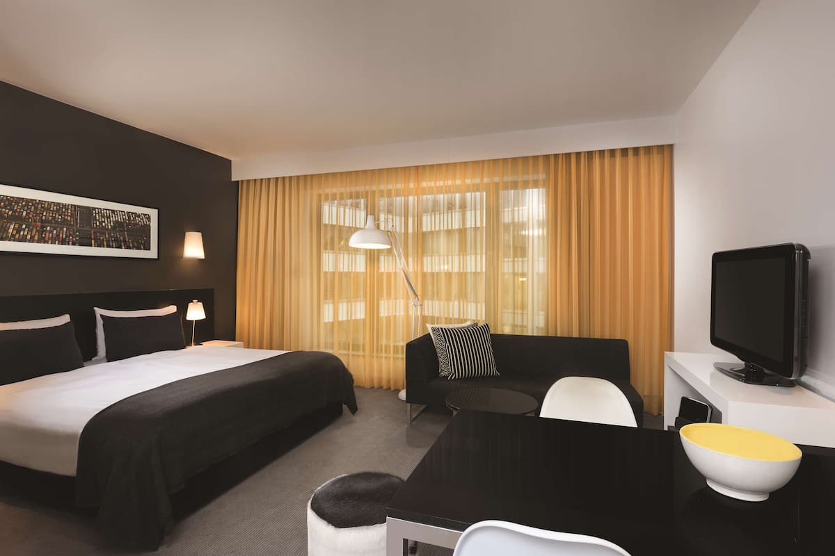 Habitació d'hotel amb un llit gran, un sofà, un escriptori amb televisió i una finestra amb cortines.
