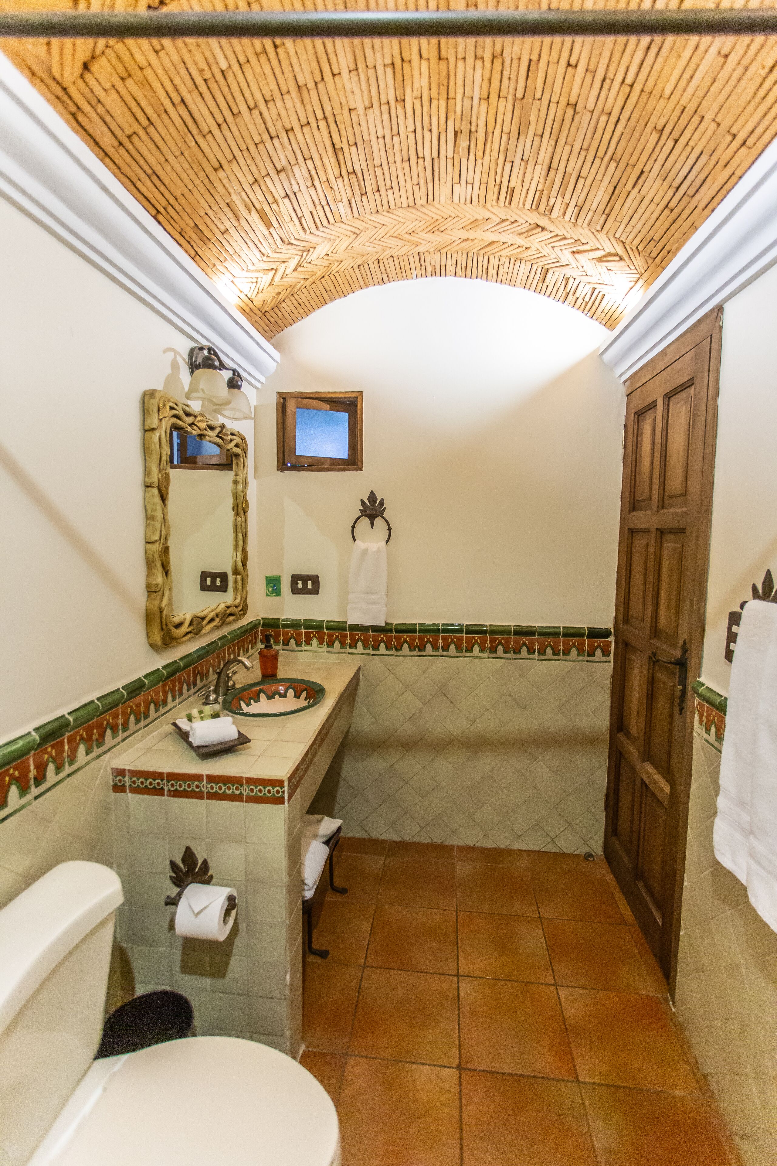 A bathroom with a bamboo ceiling, a white toilet, a sink with a decorative mirror, and a shower area.