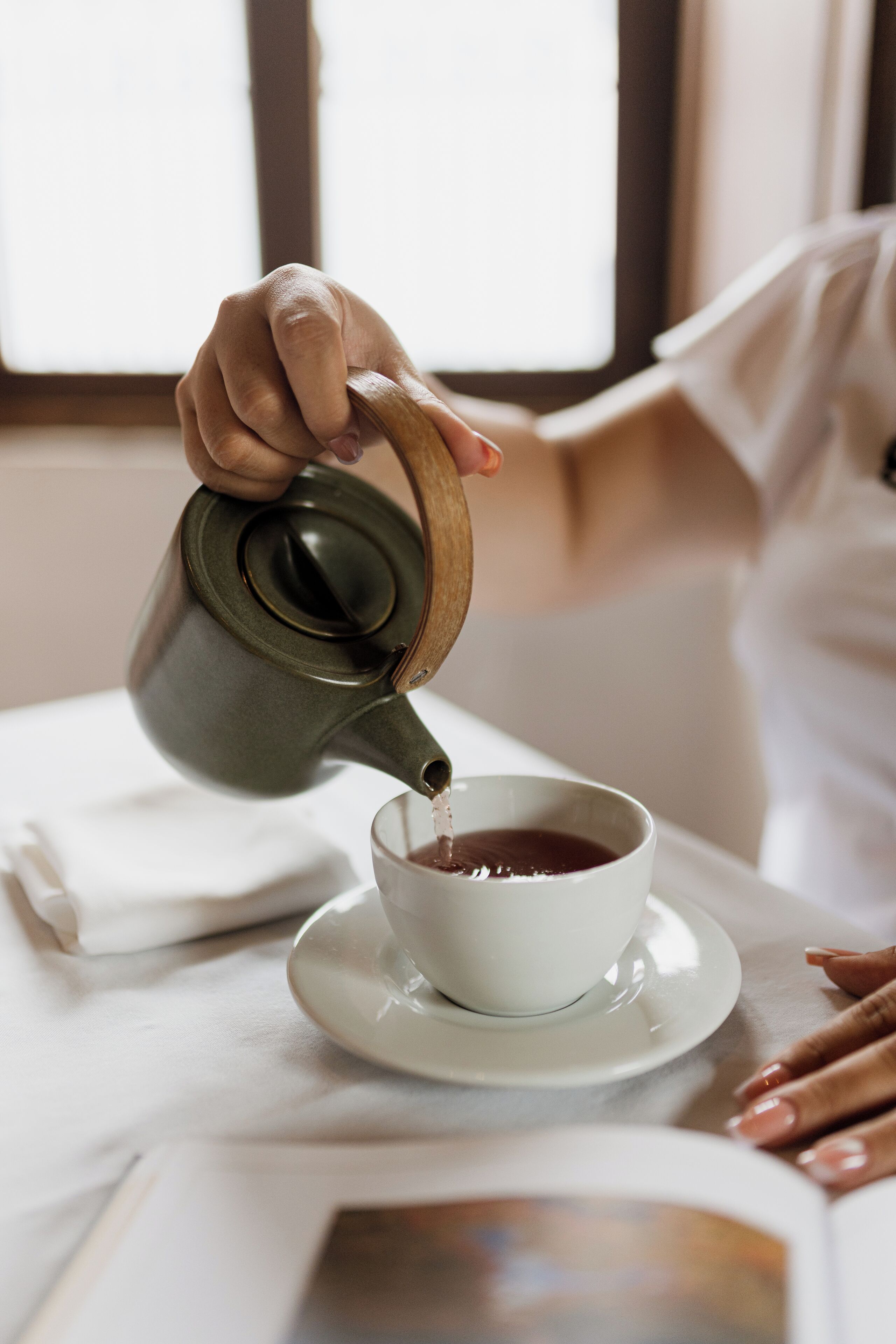 A hand pouring tea from a ceramic teapot into a white cup on a saucer.