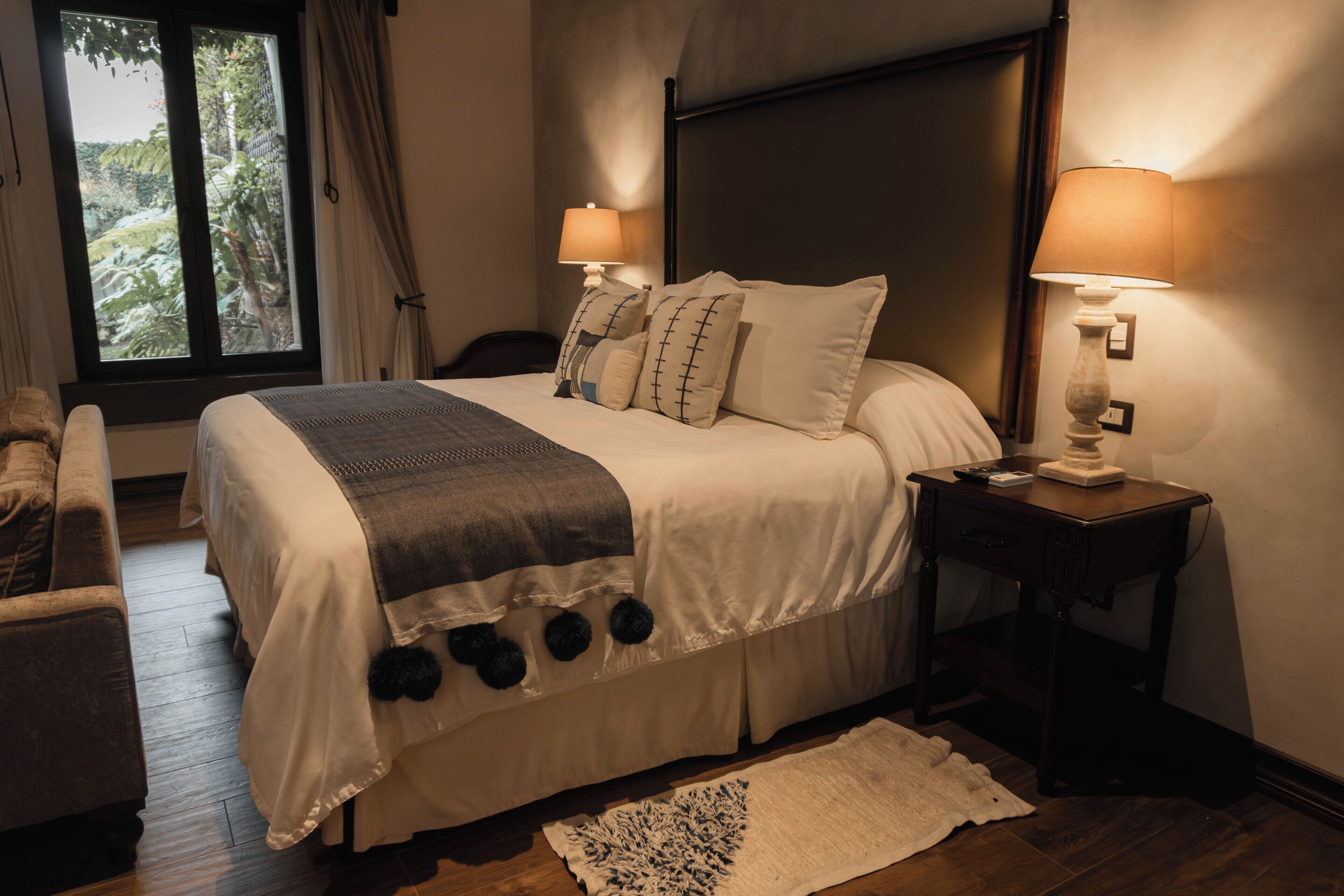 Una cama bien hecha con cabecera, dos lámparas de noche, una ventana con cortinas, piso de madera y una pequeña alfombra.
