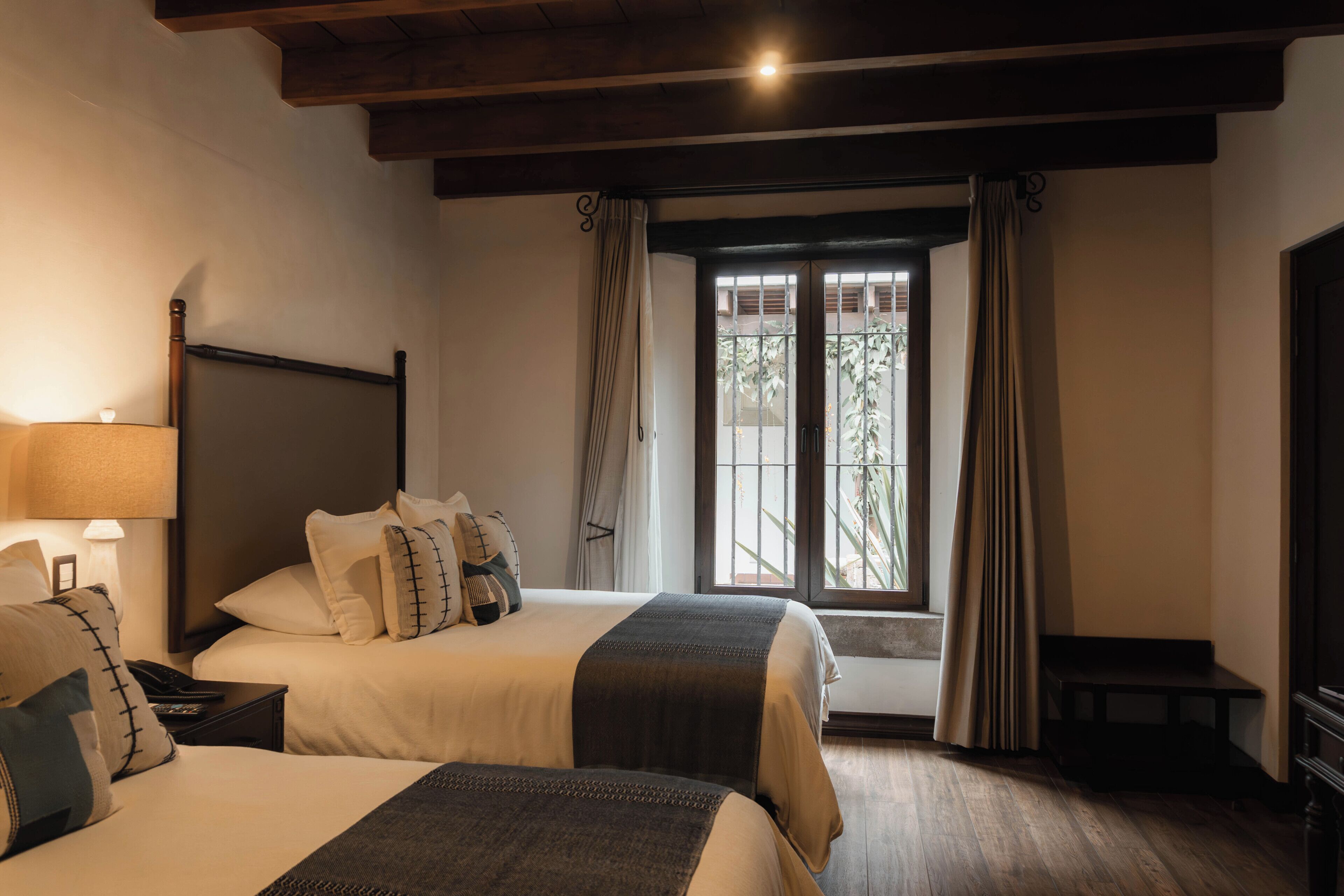 Habitación de hotel con dos camas, una mesita de noche con lámpara, un televisor y una ventana con cortinas.