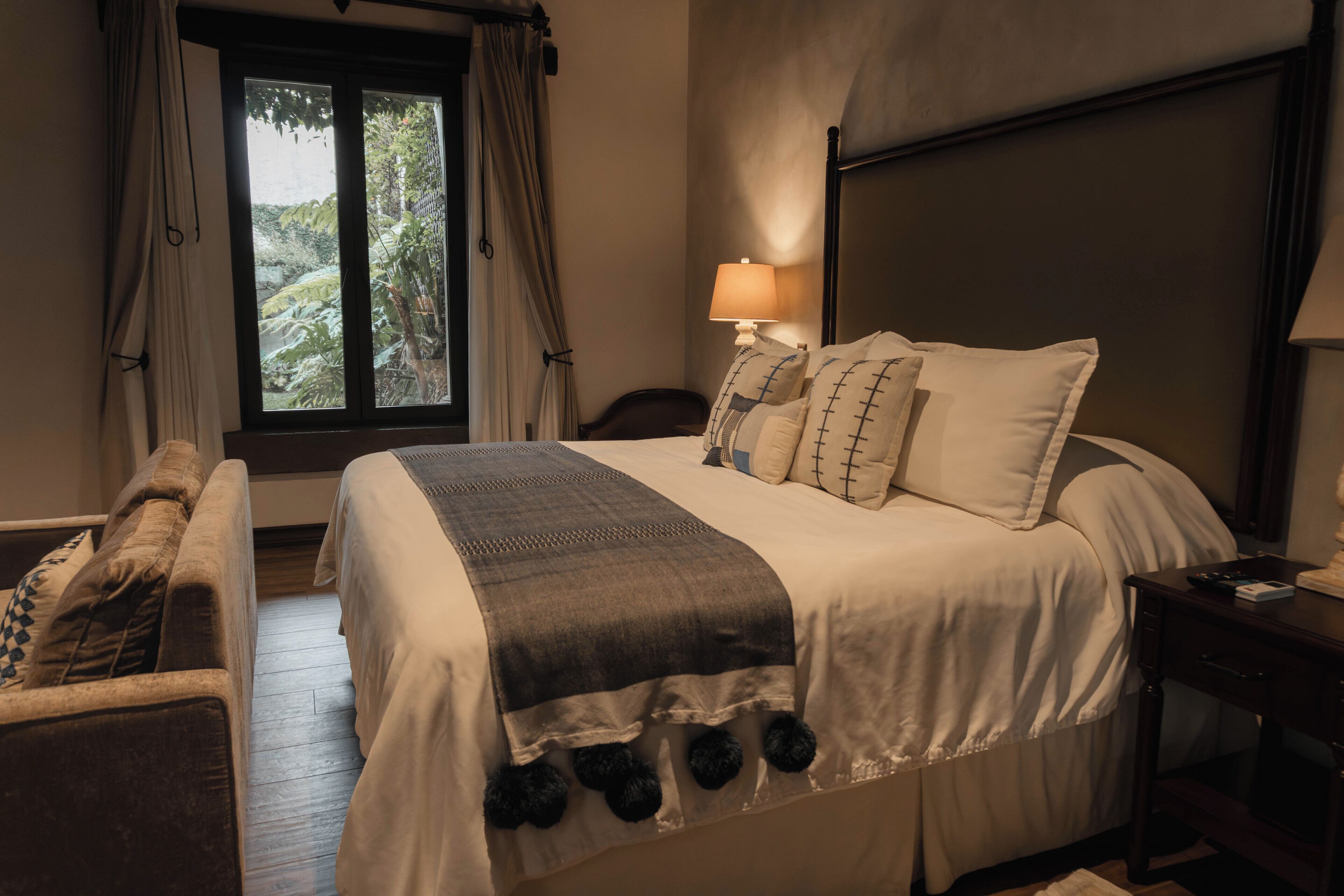 A bedroom with a bed, a bedside table, a lamp, a window with curtains, and a view of greenery outside.