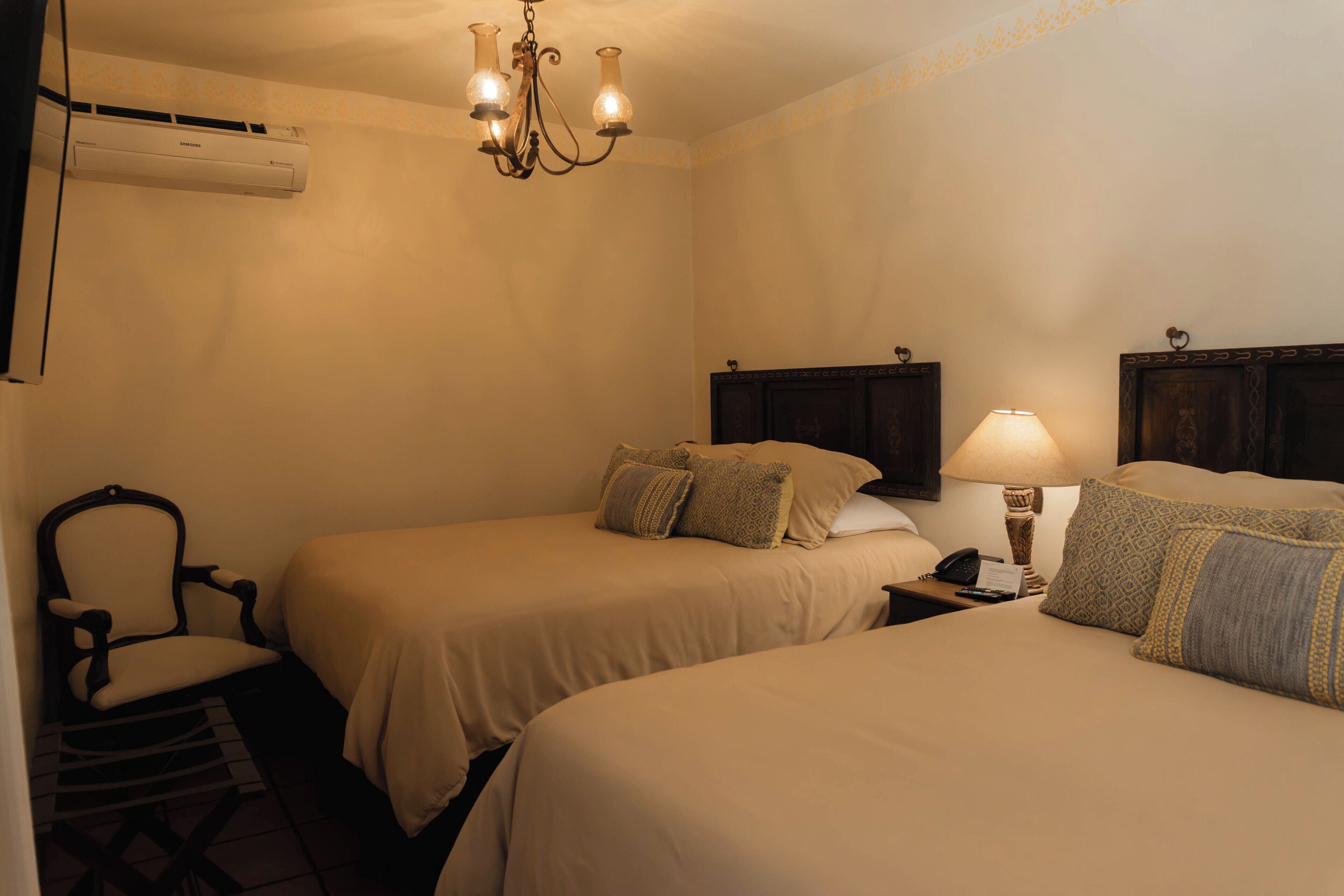 A hotel room with two beds, a chair, a nightstand with a lamp, and an air conditioning unit.