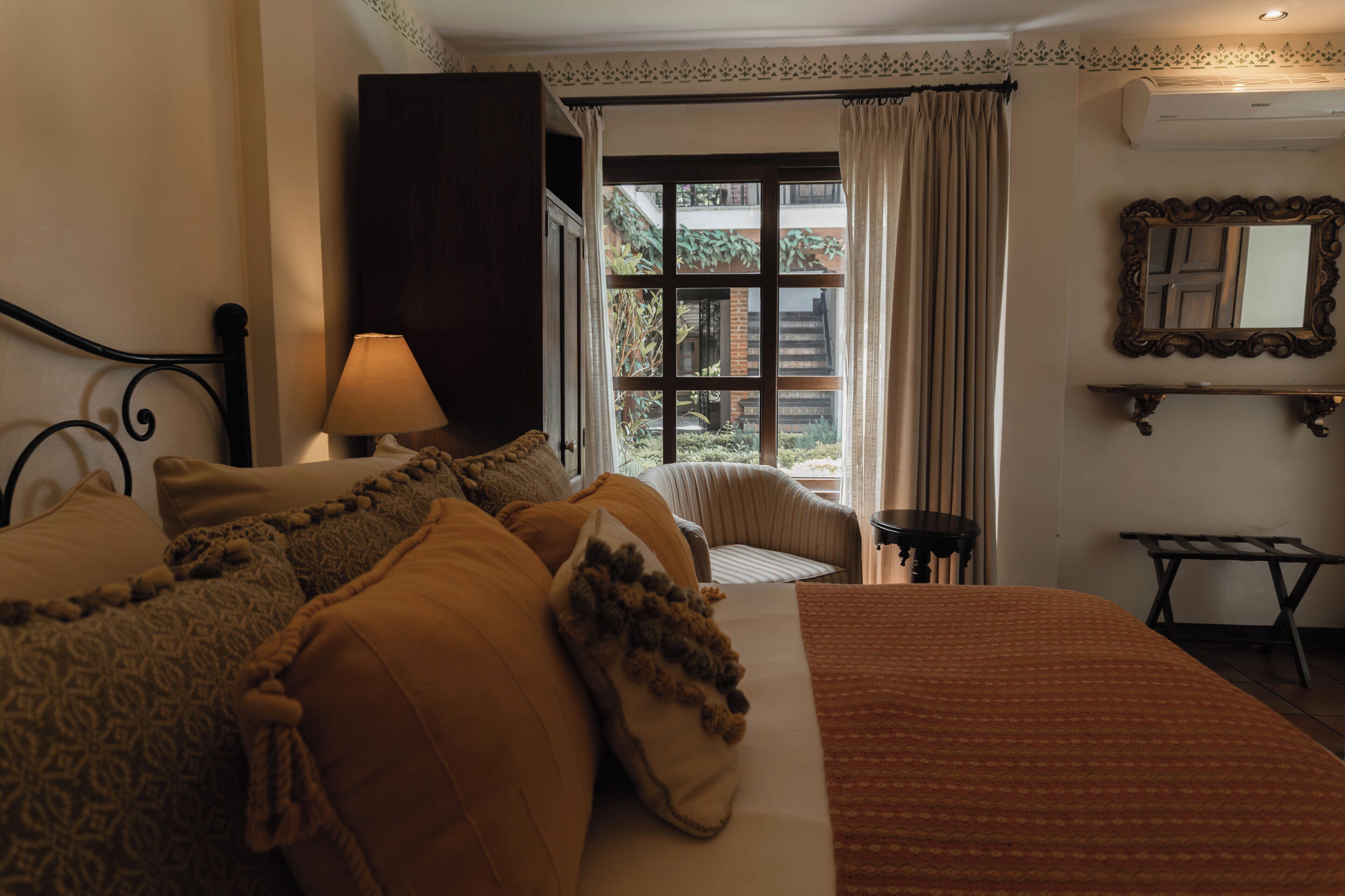 Een hotelkamer met een bed, een stoel, een lamp, een raam met gordijnen en een spiegel aan de muur.