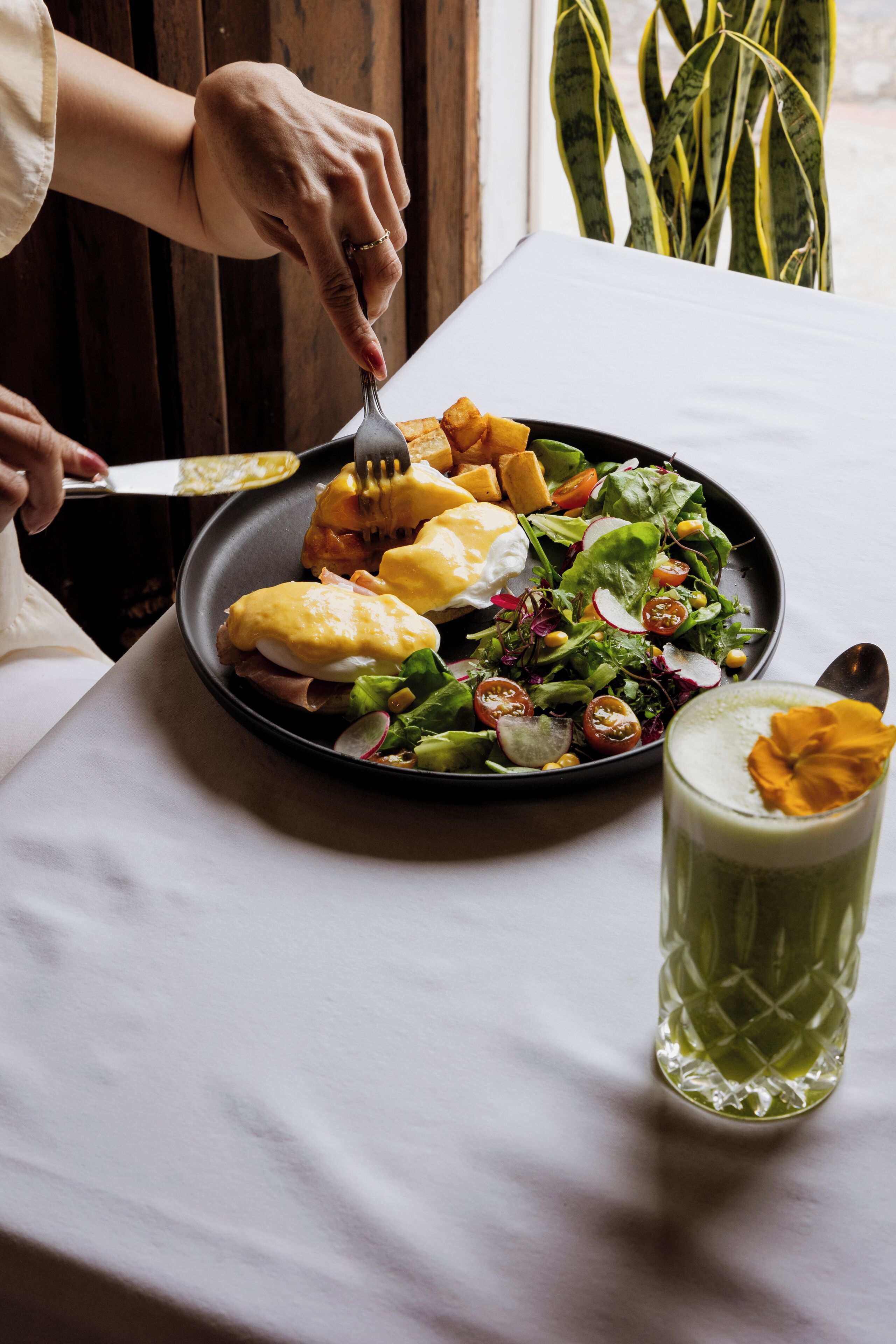 En tallrik med ägg benedict och en sallad, samt ett glas med en grön dryck på ett bord.