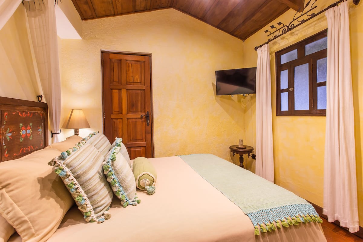 Ein Schlafzimmer mit einem Bett, einer Holztür, einem an der Wand befestigten Fernseher und einem Fenster mit Vorhängen.