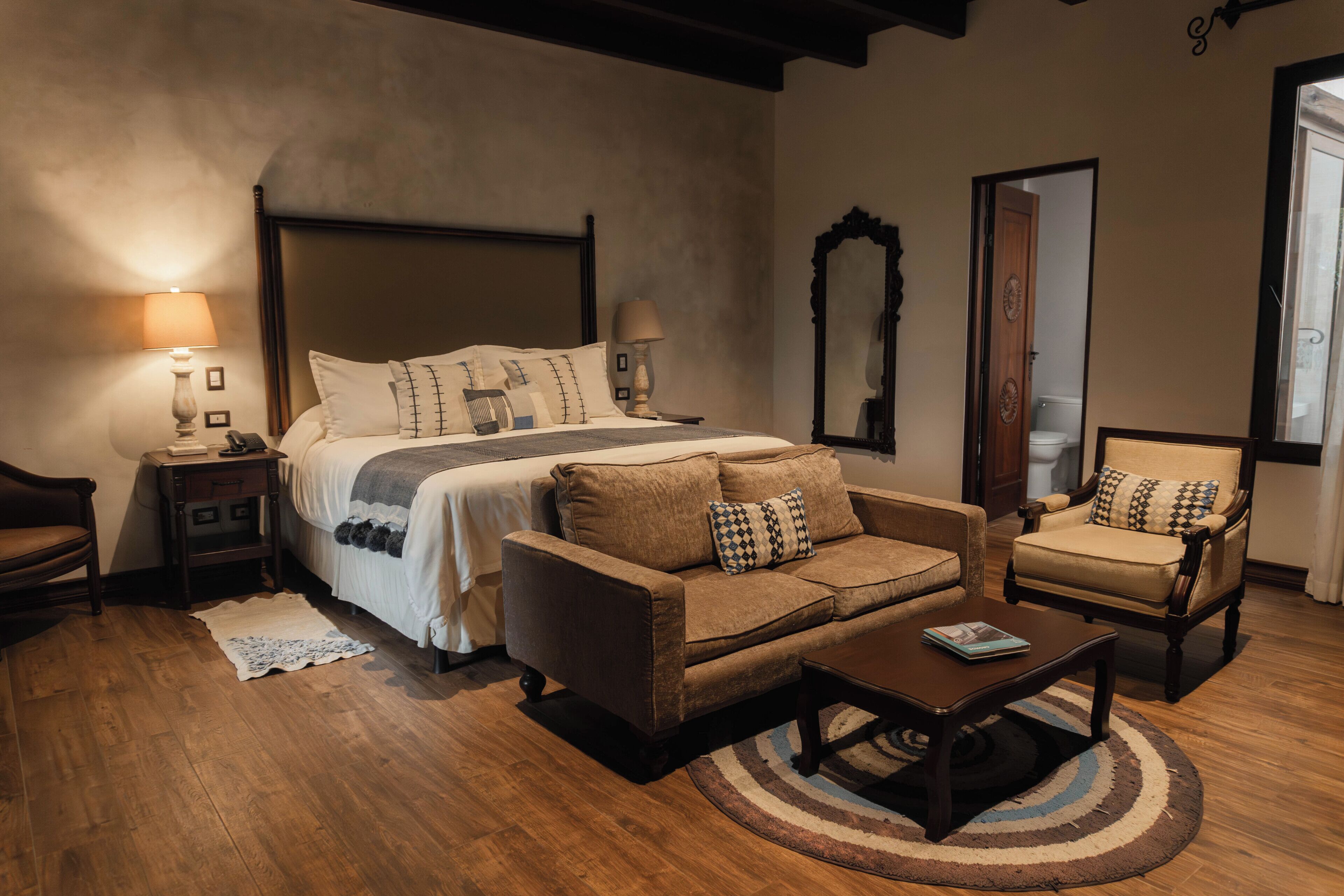 Un dormitorio con una cama grande, un sofá, una silla, una mesita de centro y una mesita de noche con lámpara.