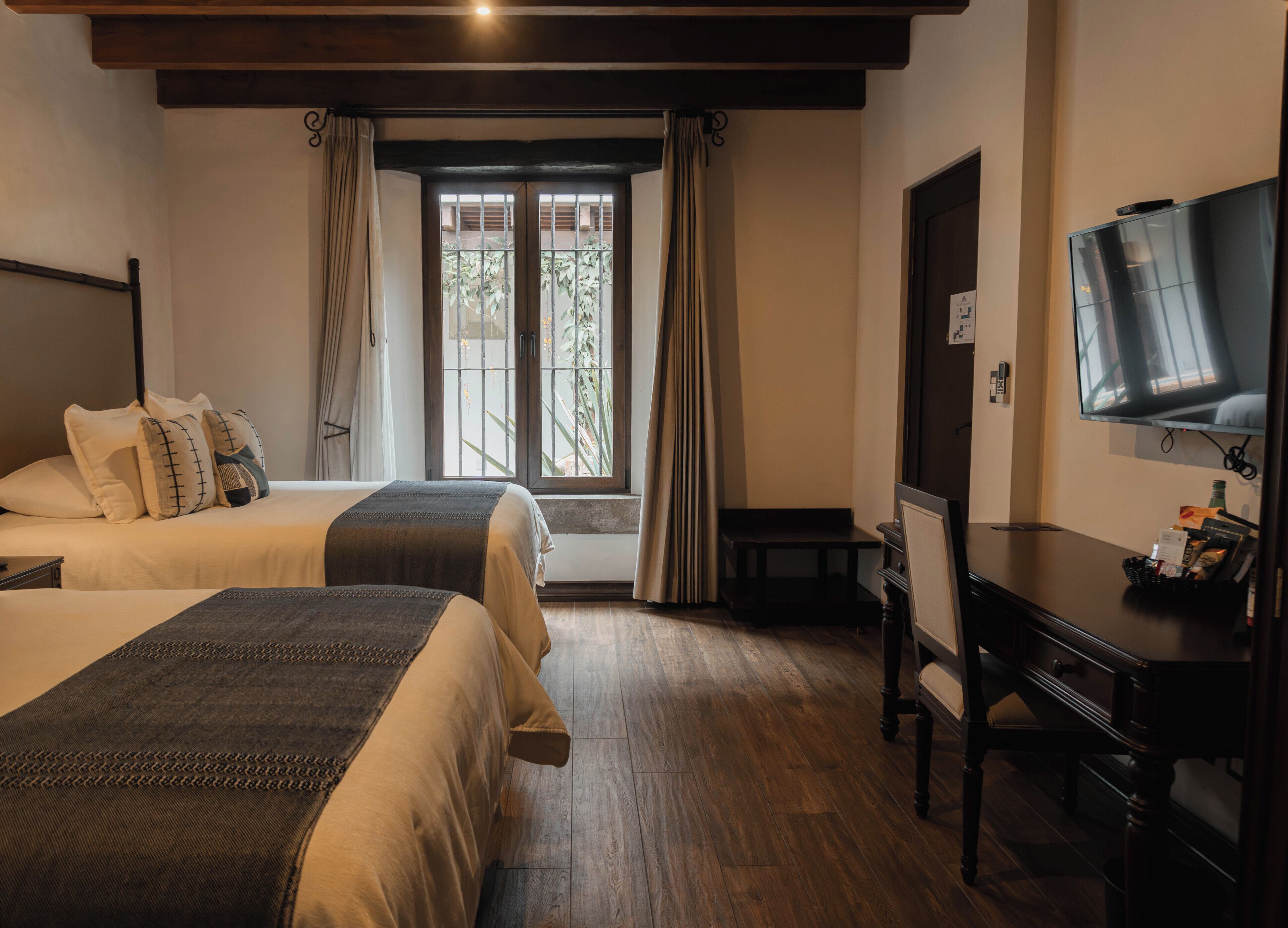 A hotel room with two beds, a desk, a television, and a window with curtains.