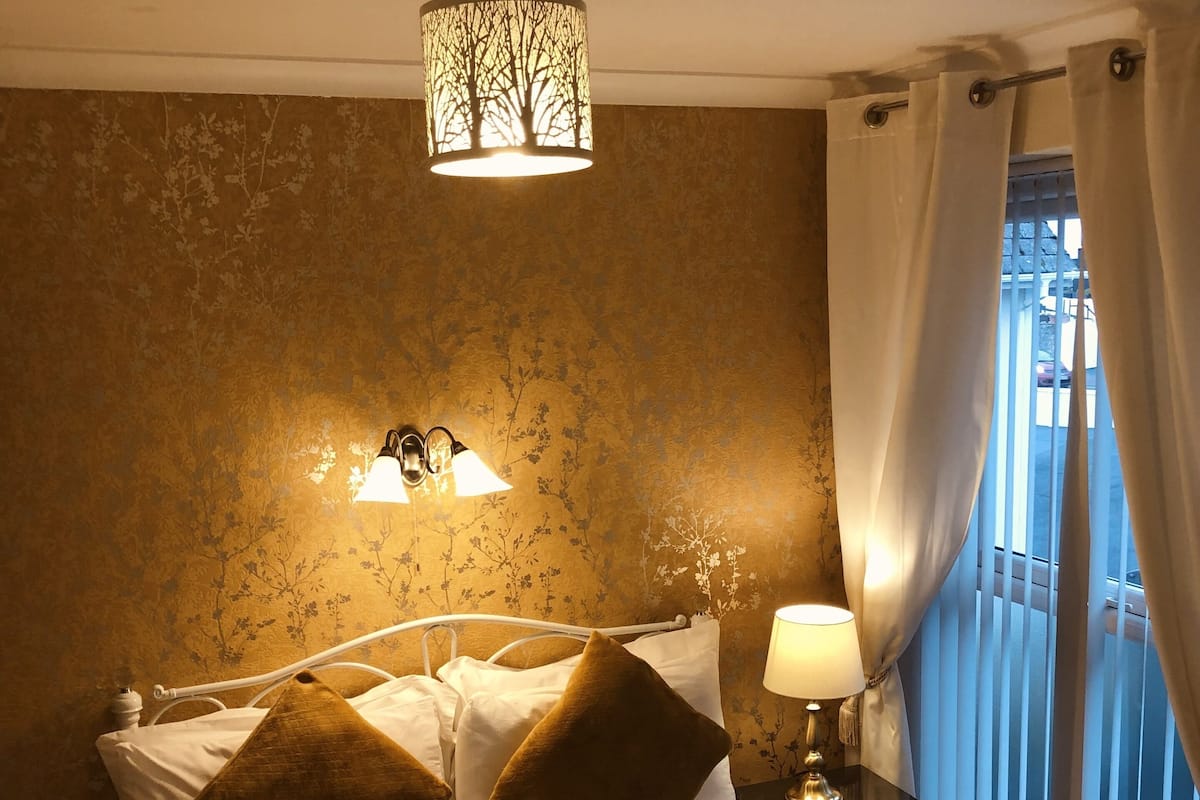 A hotel room with a bed, a nightstand with a lamp, a window with curtains, and a wall with a floral pattern.