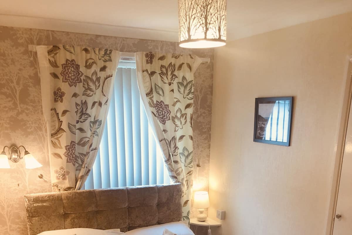 A neatly made bed with floral-patterned pillows and a blue bag, a bedside lamp, and a window with floral curtains.