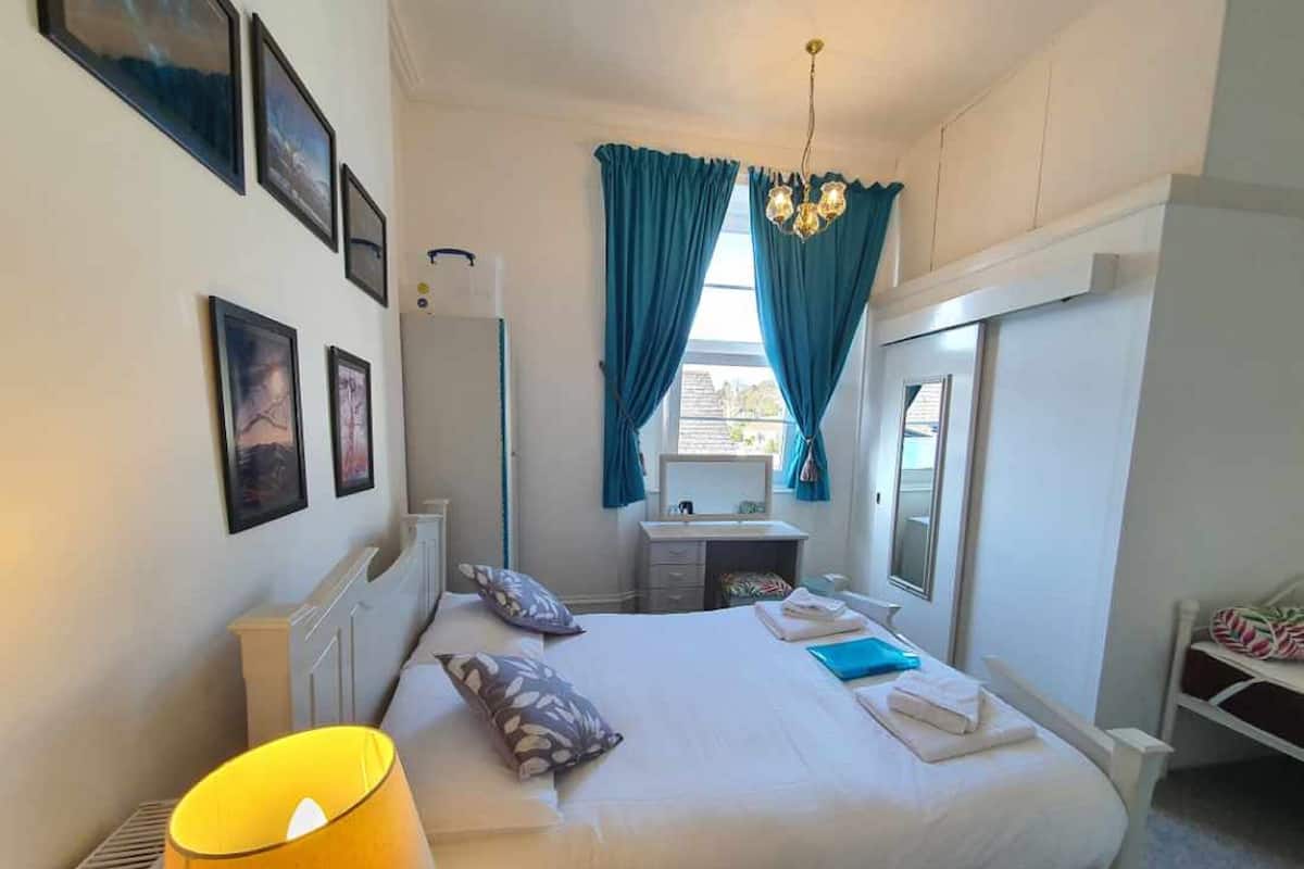 A bedroom with a bed, a bedside lamp, a desk with a chair, a window with curtains, and wall-mounted pictures.