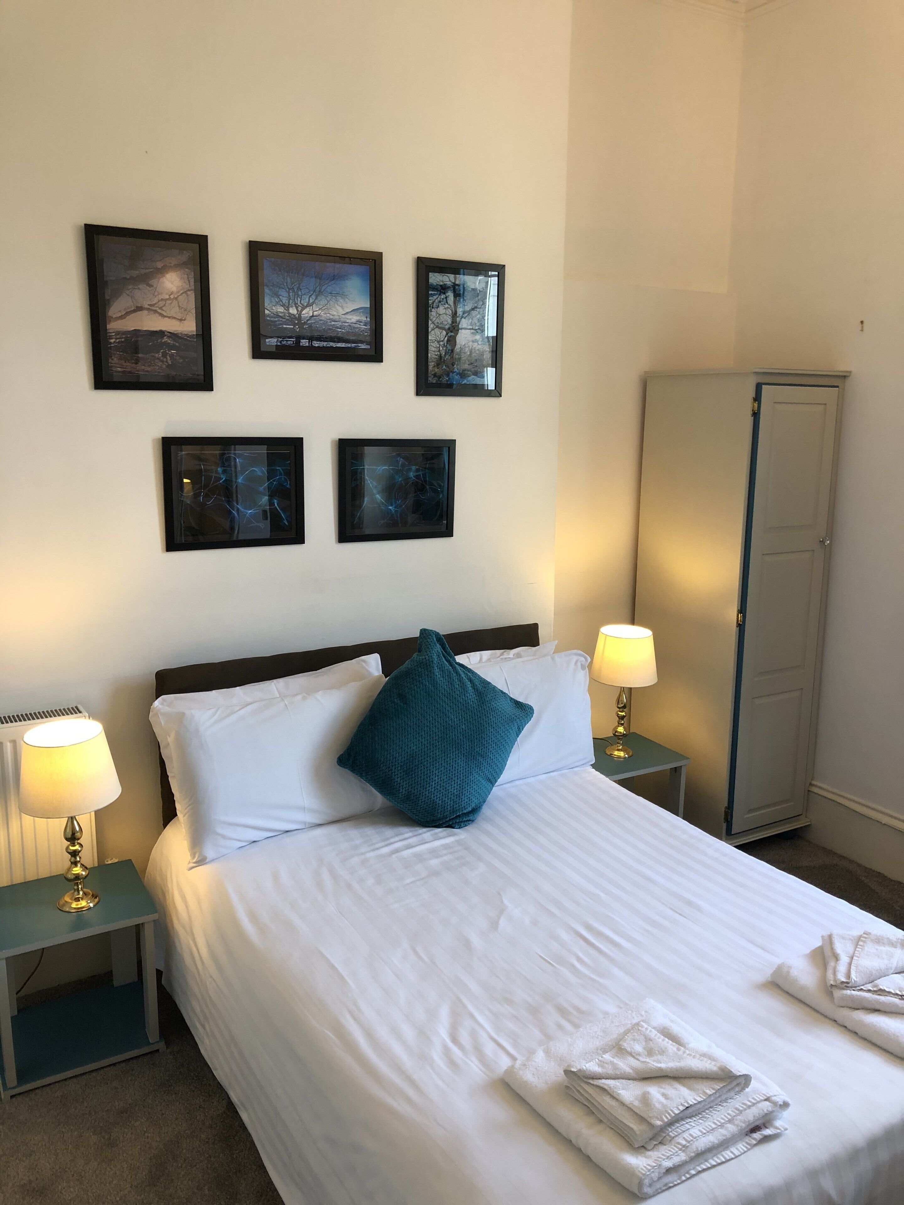 A neatly made bed with white linens and a teal pillow, flanked by two bedside tables with lamps, in a room with a wardrobe and framed pictures on the wall.