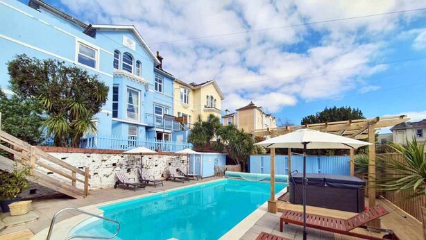 A swimming pool with lounge chairs and umbrellas, surrounded by a stone wall and a wooden deck. - The Purple Tree Guest House (Torquay)