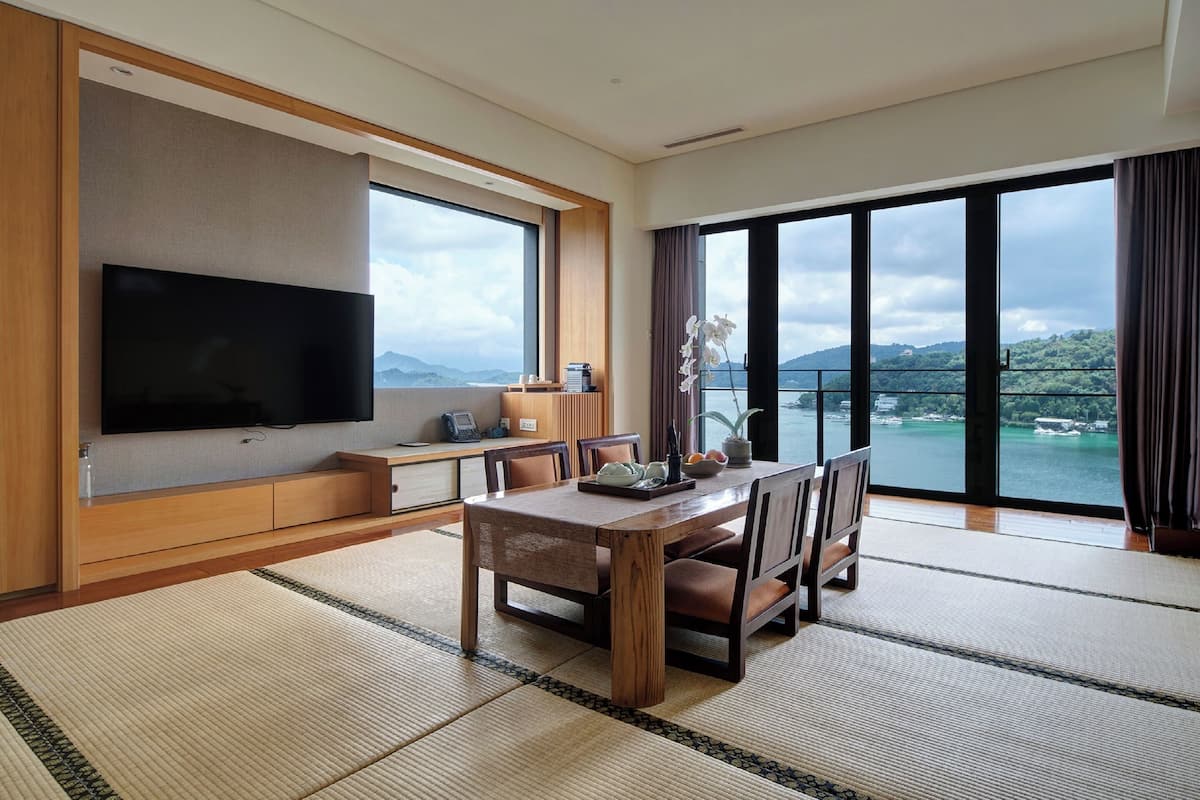 Habitación de hotel moderna con un televisor de pantalla plana grande, una mesa de comedor de madera y vistas a un cuerpo de agua y montañas.