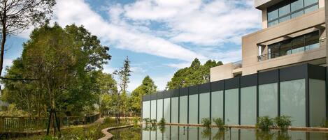 A modern building with large glass windows, a reflection in a water feature, and surrounding greenery.