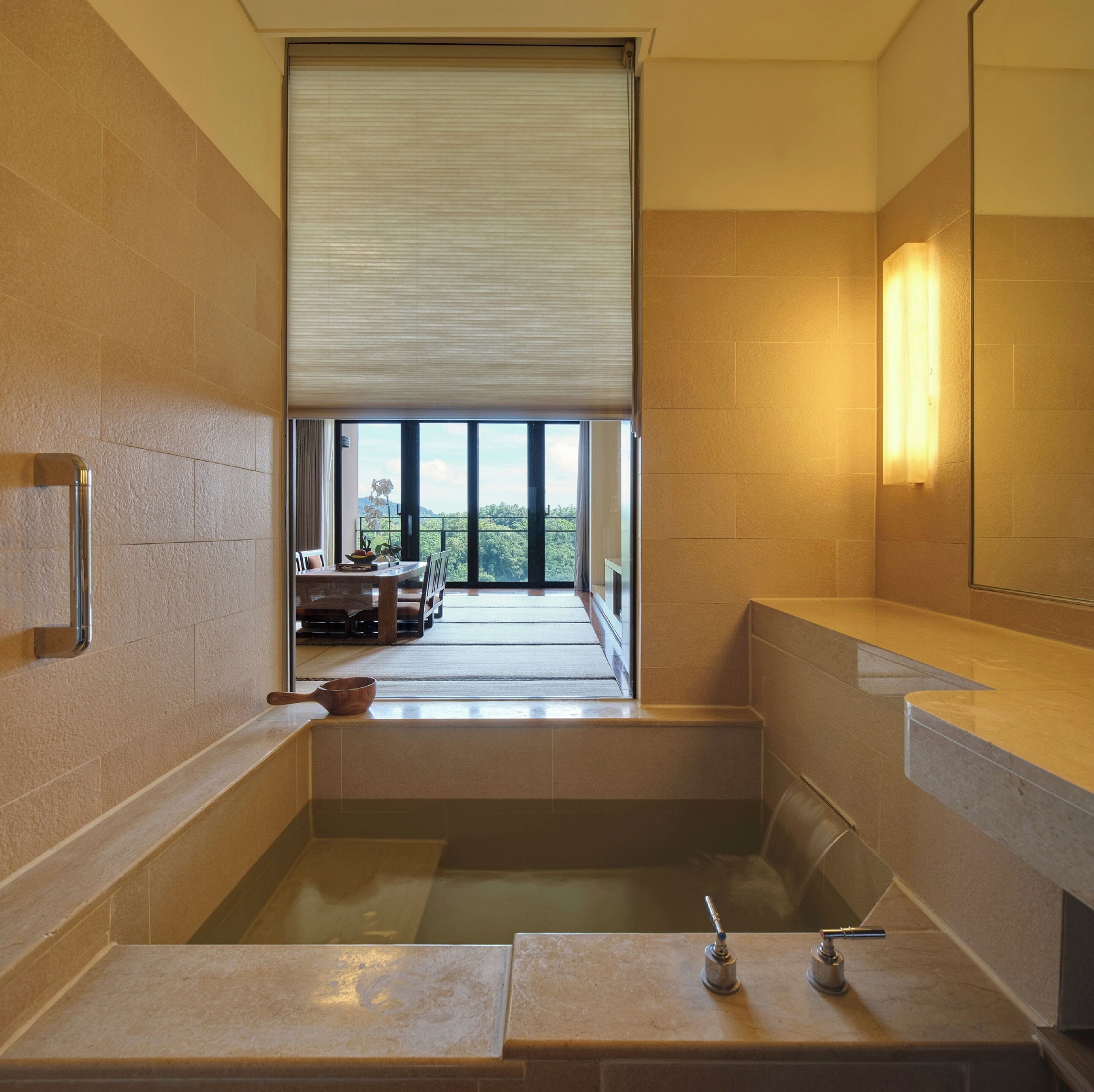 A modern bathroom with a large bathtub, a toilet, and a sink. There is a window with a view of a balcony and greenery outside.