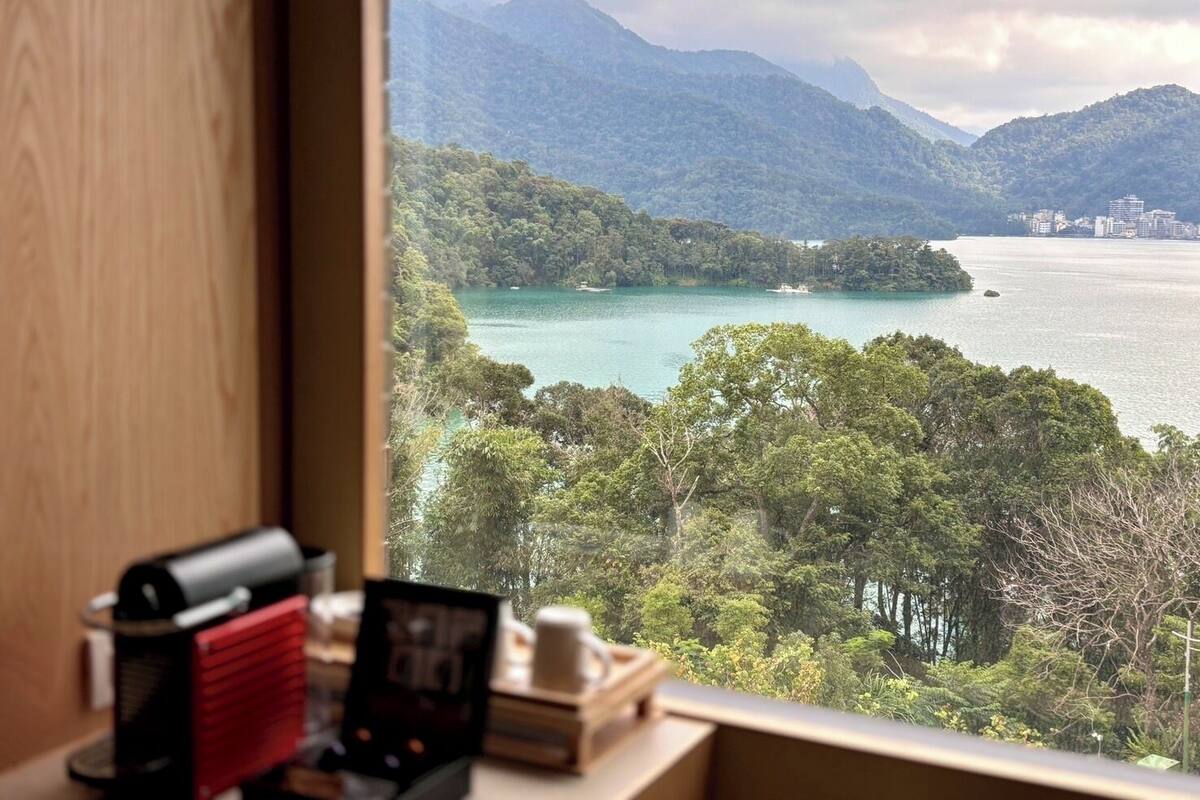 Vista desde una ventana sobre una bahía con montañas, una pequeña isla y un horizonte urbano distante.