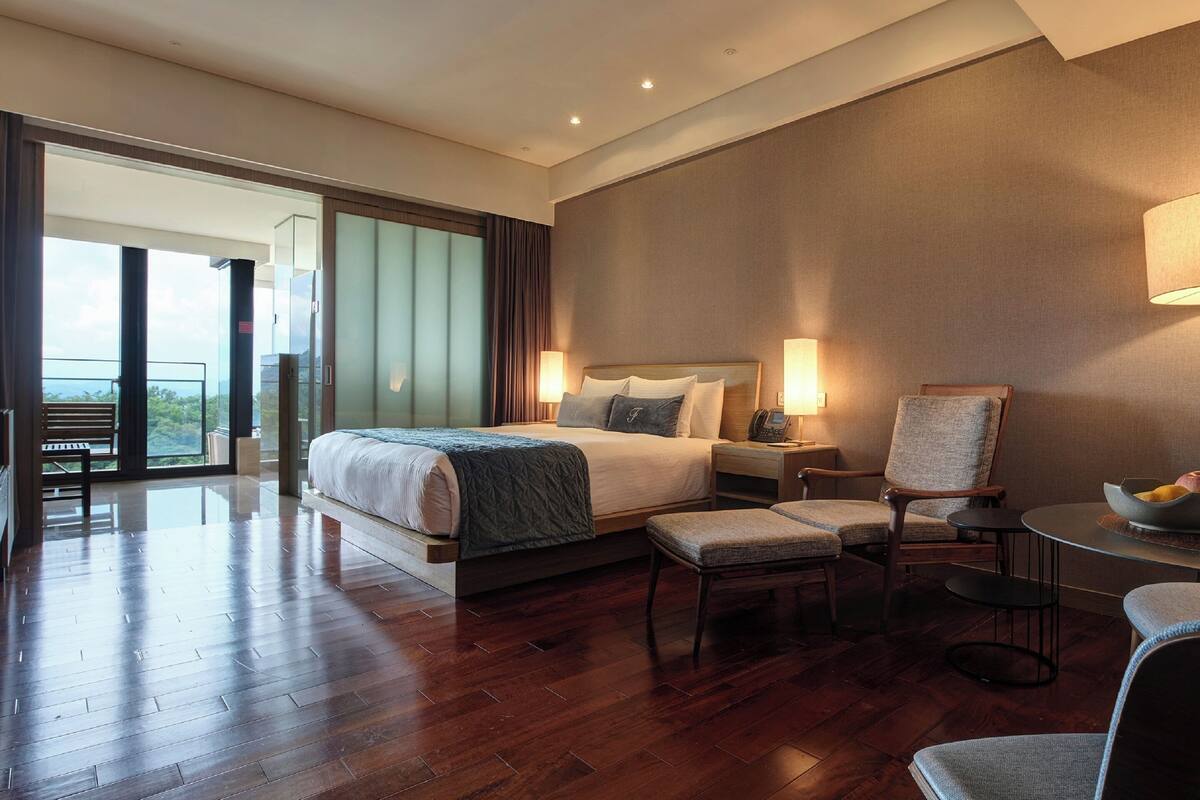Amplia habitación de hotel con una cama grande, zona de estar con un sillón y reposapiés, una mesita con un frutero y un balcón con vistas.
