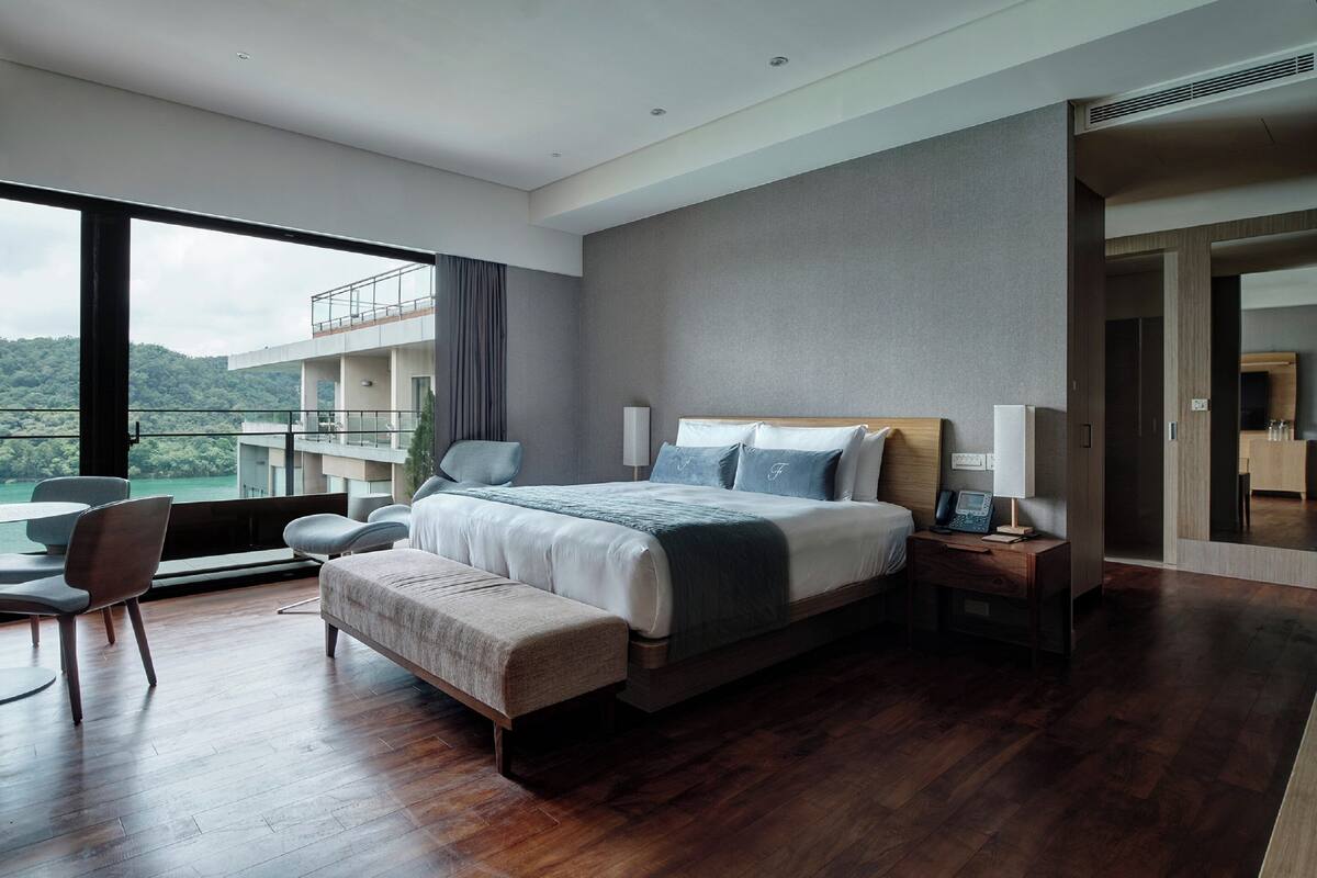 Habitación de hotel moderna con una cama grande, una zona de estar con un banco, un escritorio con lámpara y vistas a un balcón y montañas.